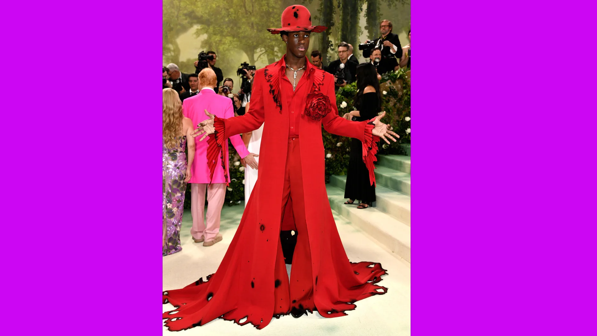 A photograph of Wisdom Kaye in a red dress coat and suit with hat and the suit has burn marks around the hem and collar and on the hat. There are press photographers behind him.