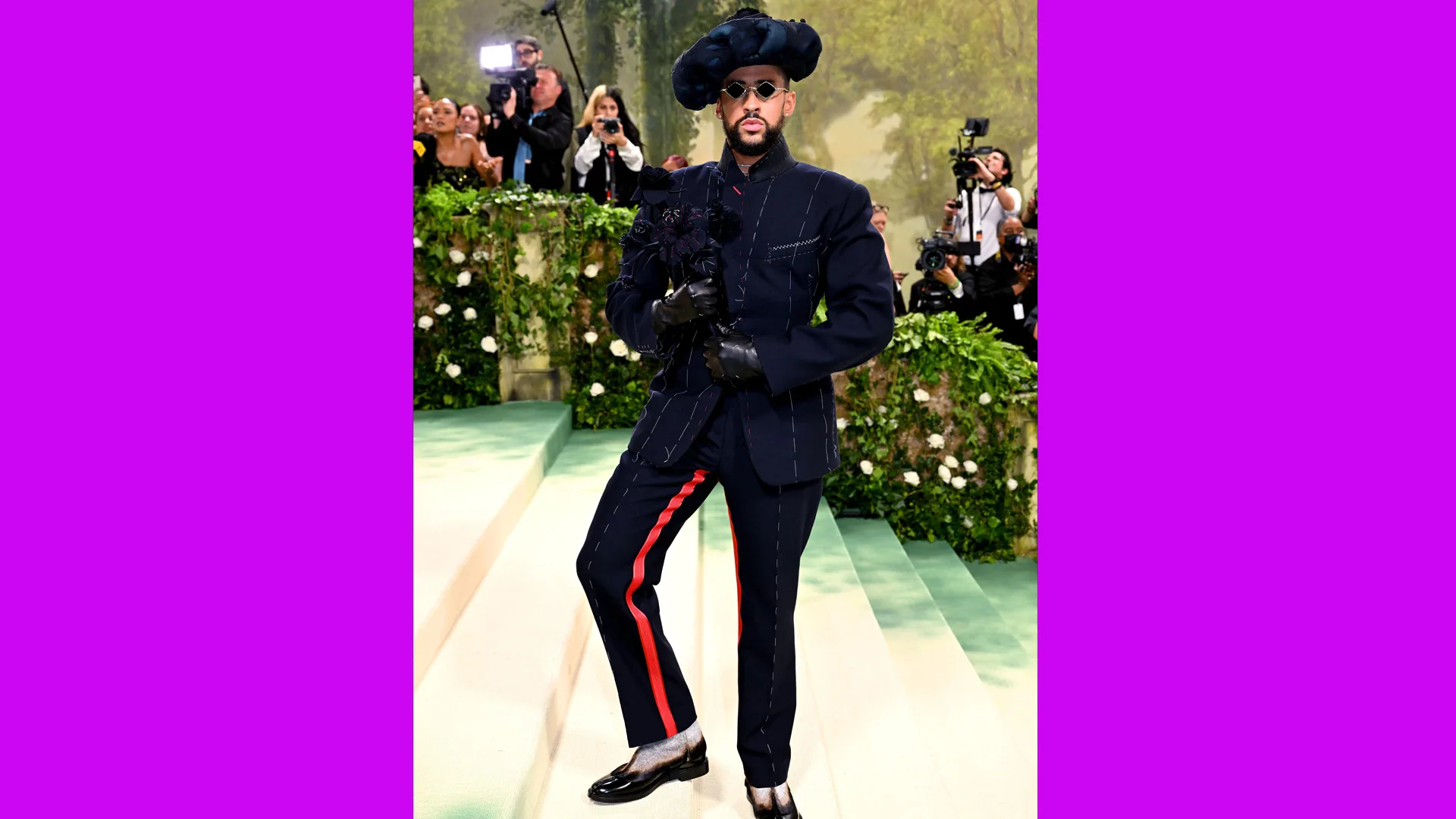 A photograph of Bad Bunny at the Met Gala 2024 wearing a black suit with a black hat on a white staircase with press photographers behind