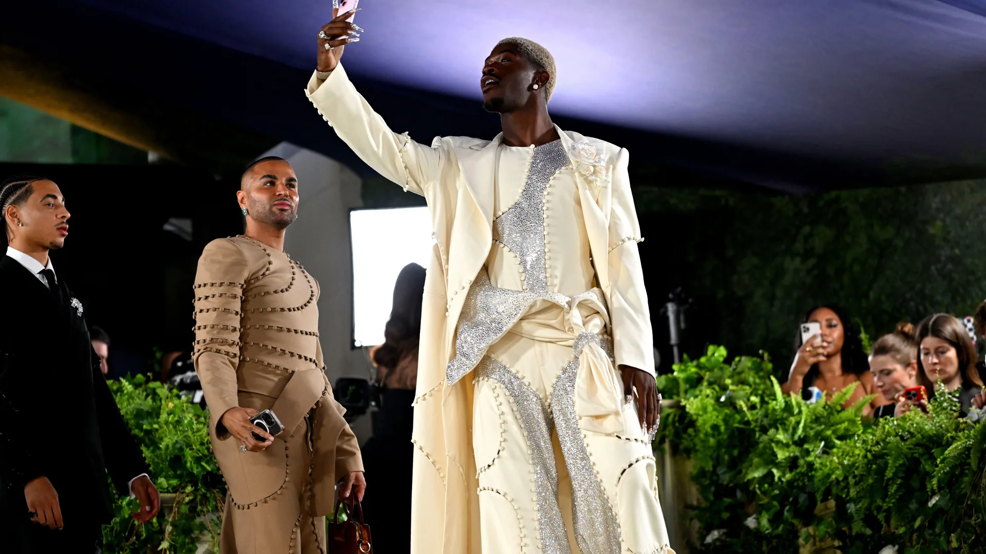A photograph of Lil Nas X at the Met Gala 2024 wearing a white and silver suit taking a selfie with guests and photographers behind him with a bush