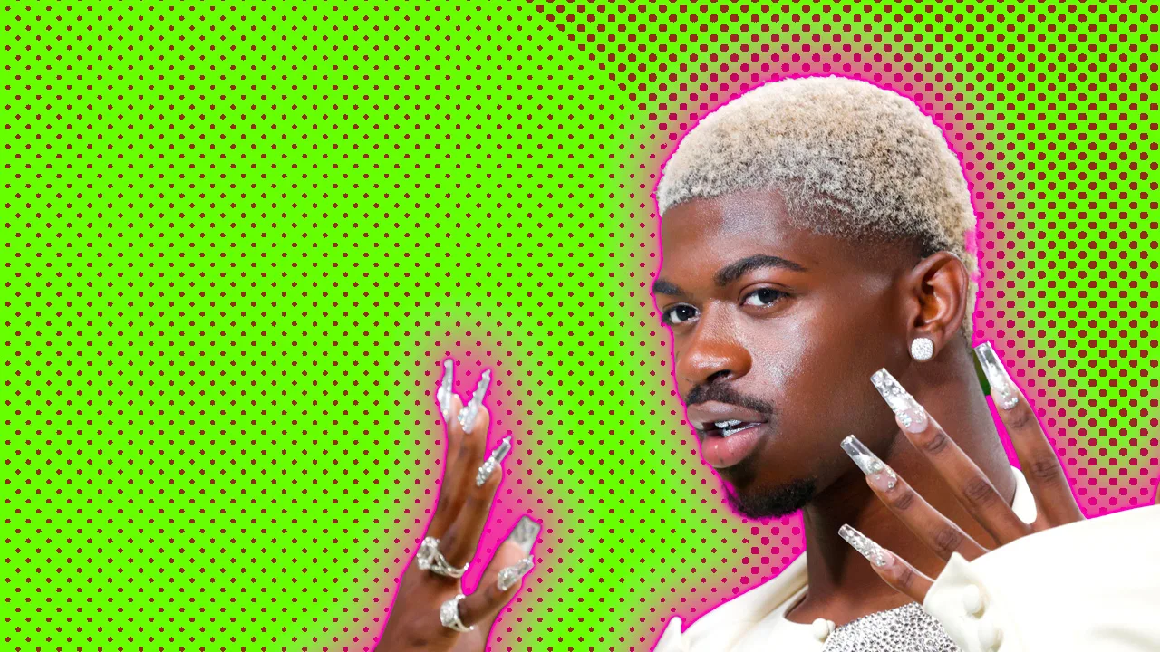 A photograph of Lil Nas X holding up his hands with big silver fake nails against a green dotted background with a pink halo