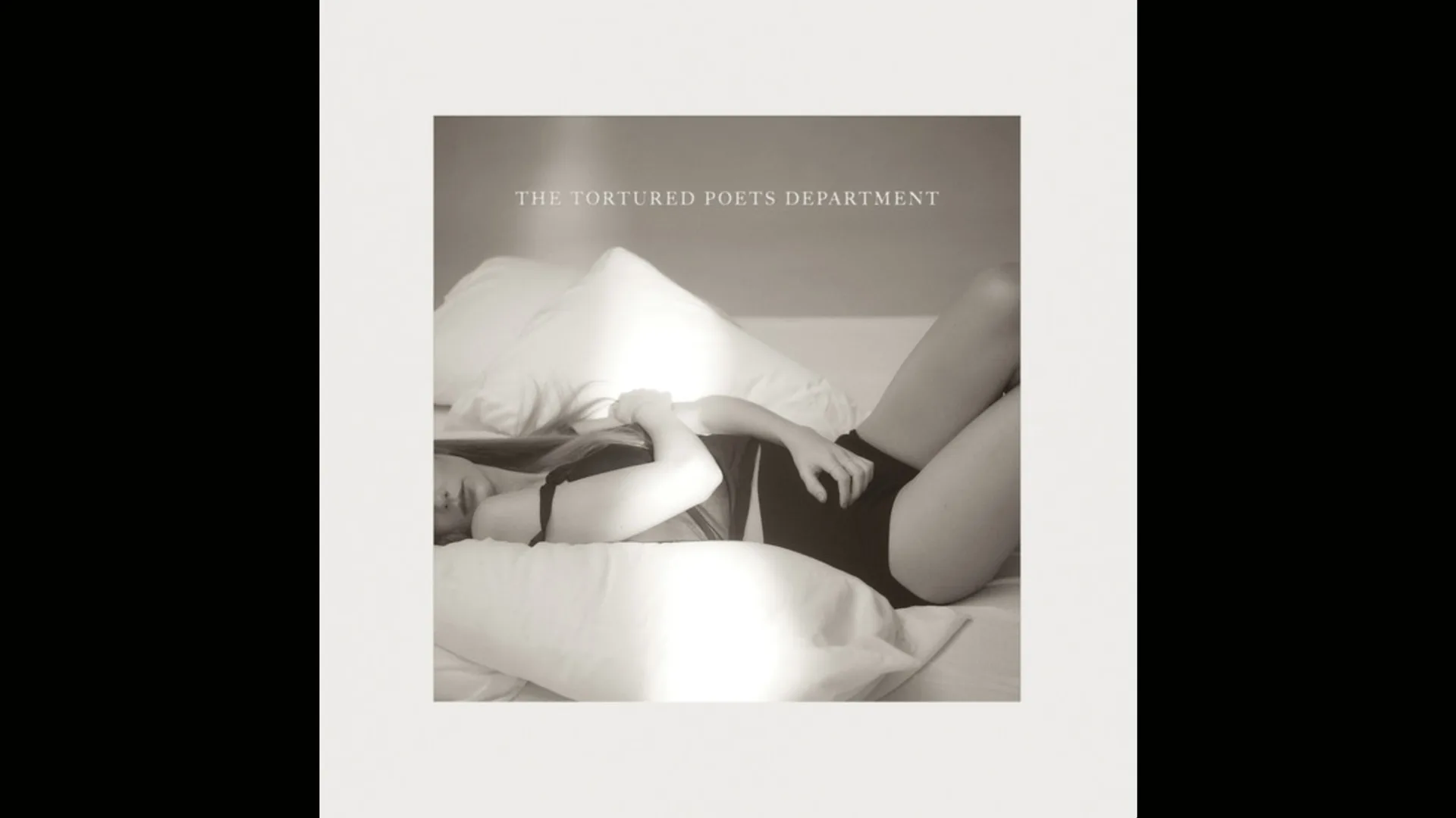 Taylor Swift's album cover for THE TORTURED POETS DEPARTMENT showing the singer lying on a bed on big white pillows wearing a black top and shorts cuddling herself. The image is in black and white and the text of the album title is above her in white.