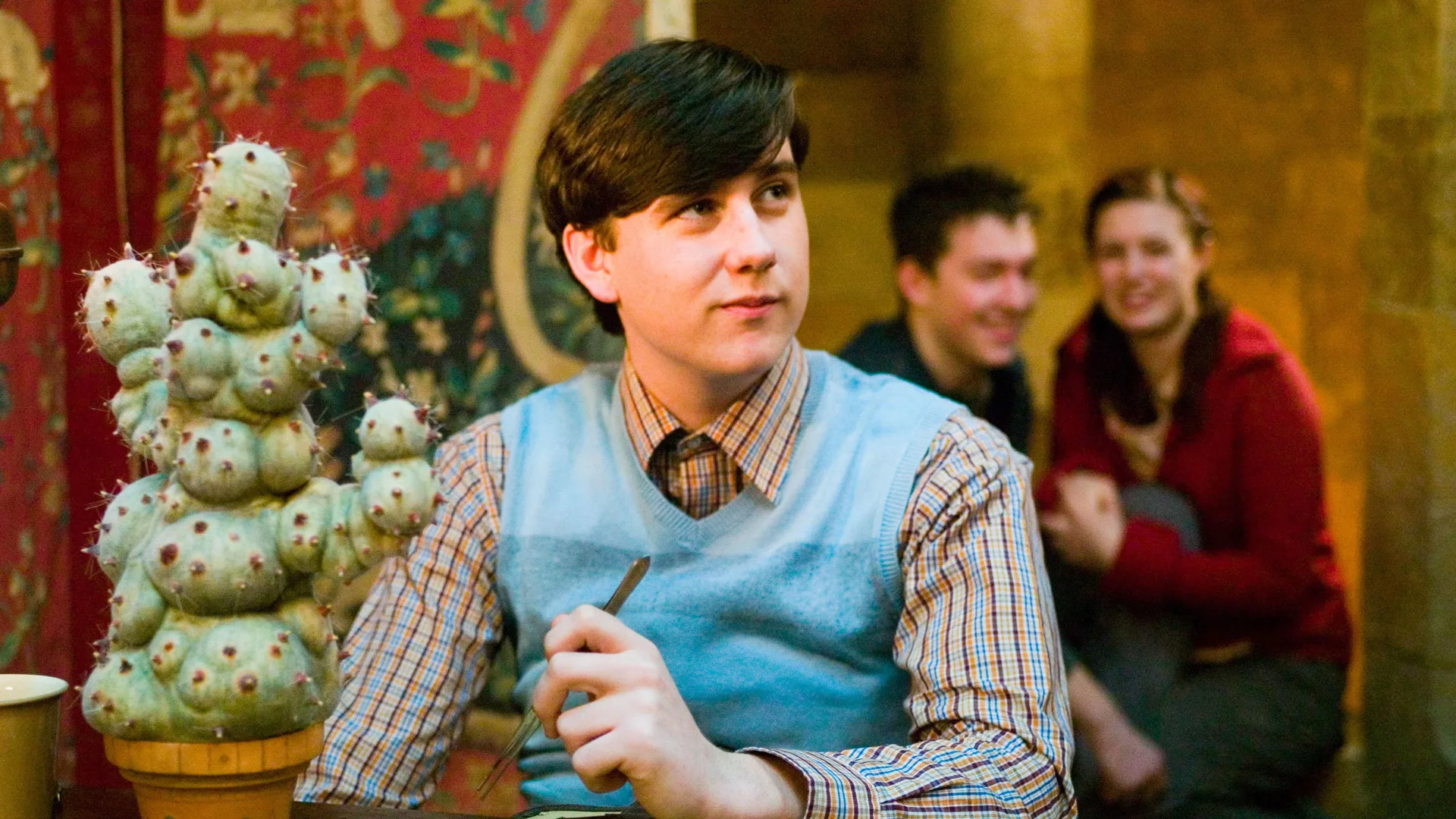 A still from Harry Potter showing Neville Longbottom writing something in a blue jumper with a smile on his face. Behind him are two people smiling.