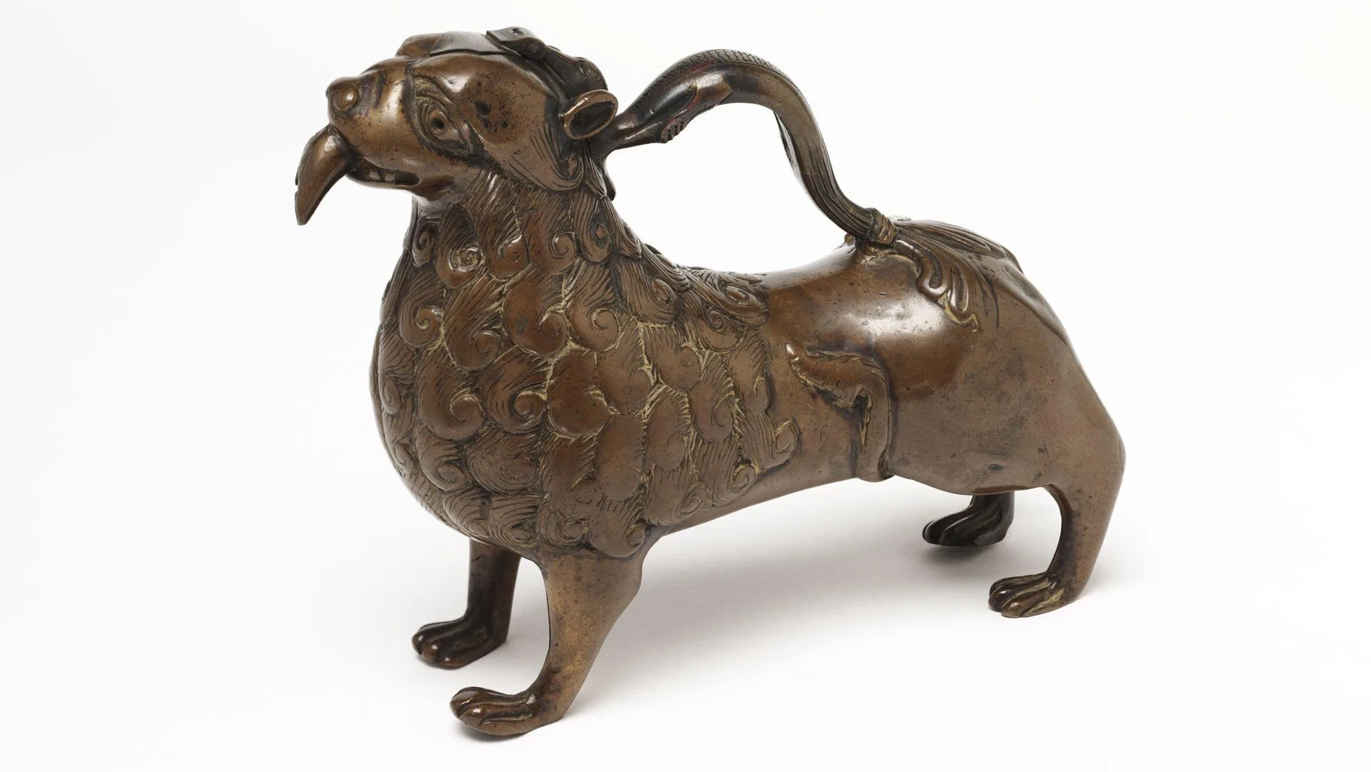 A photo of bronze jug in the shape of a lion with it's tail as the handle. It is stood on all fours with it's tongue sticking out against a white background.