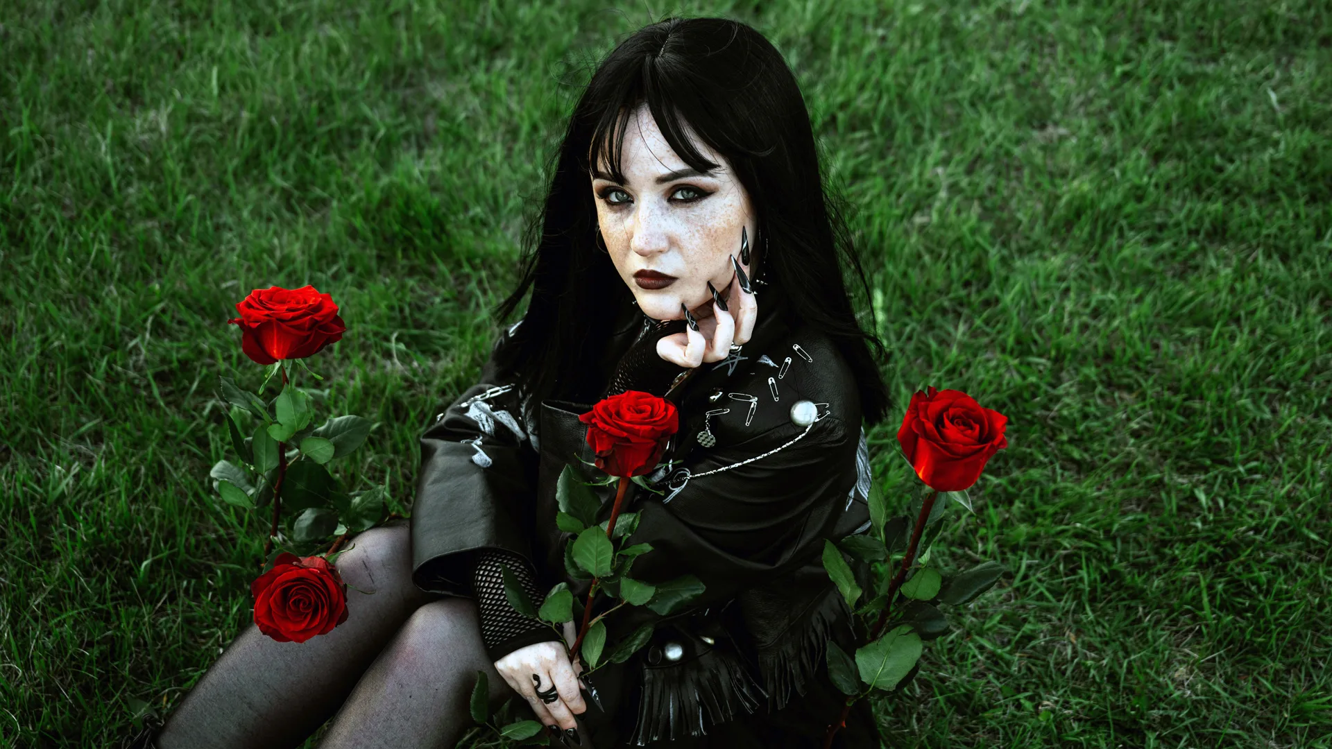 A photo of a girl wearing gothic clothing and makeup sat in grass surrounded by red roses. She has one hand to her face with black nails and is looking directly at the camera.