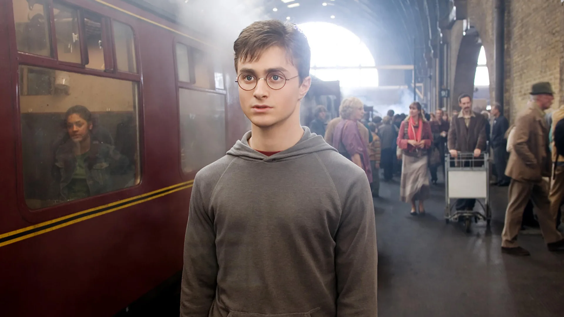 A photo of Daniel Radcliffe in Harry Potter standing at a train station with a concerned look on his face as crowds walk behind him.
