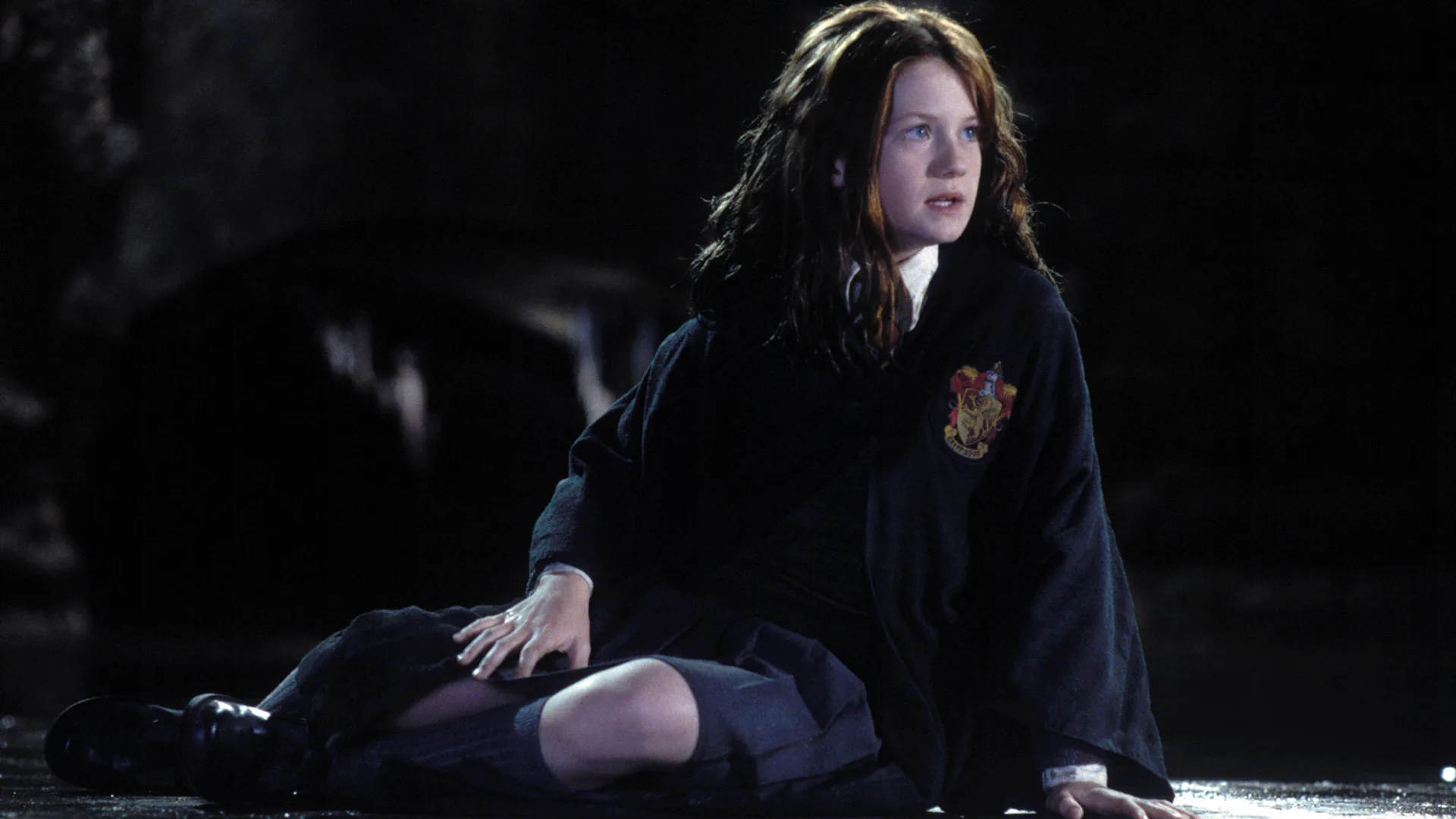 A photo of Ginny Weasley in Harry Potter sat on the floor looking distressed to something to the side - she is surrounded by darkness.