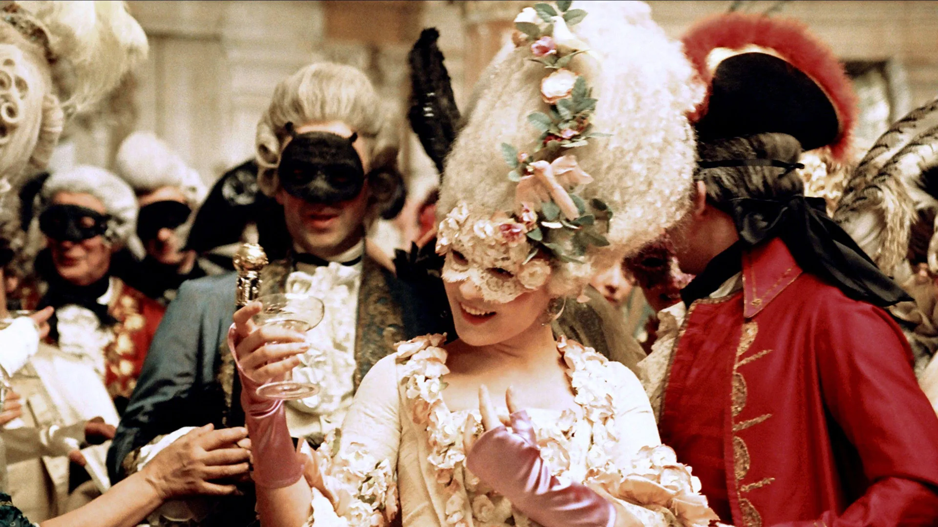 A still from the Sofia Coppola move Marie Antoinette (2006) showing Kirsten Dunst as Marie Antoinette wearing a giant white wig decorated with flowers and ribbons and a white mask smiling to her guests who are also wearing masks and wigs in a party setting.