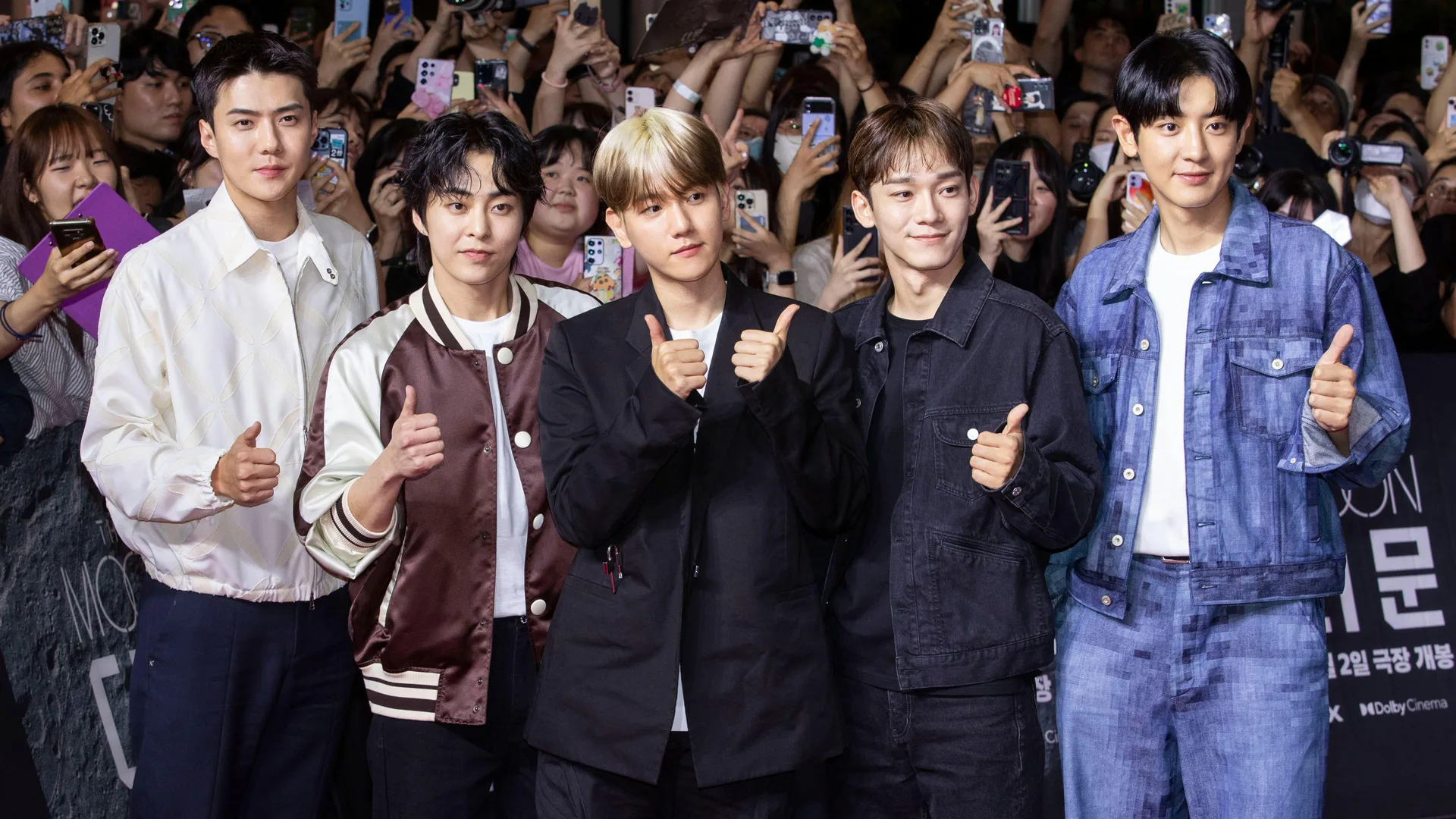 An image showing five members of the K-pop band EXO stood in a crowd looking at cameras with their thumbs up smiling.