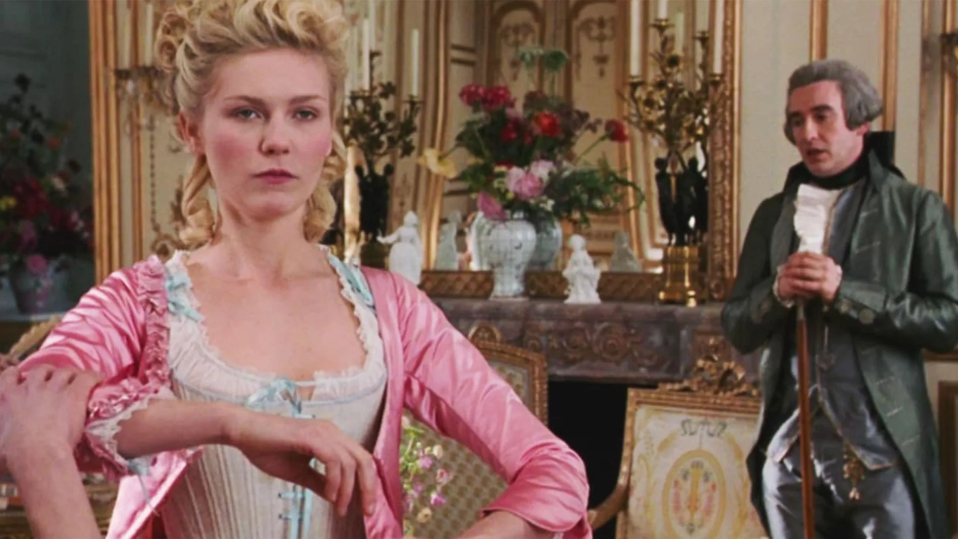 A still from the Sofia Coppola move Marie Antoinette (2006) showing Kirsten Dunst as Marie Antoinette being dressed in a pink gown as her teacher stands behind her. She has a stern expression.