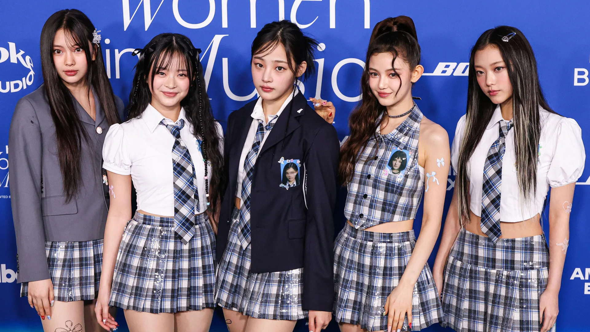 A photo of the k pop band NewJeans showing the five members stood in a line smiling wearing plaid school uniform like outfits.
