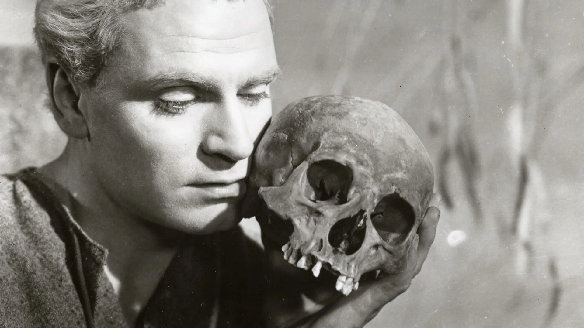 A still from Hamlet (1948) starring Lawrence Olivier showing him holding a skull close to his face and gazing at it against a grey background.