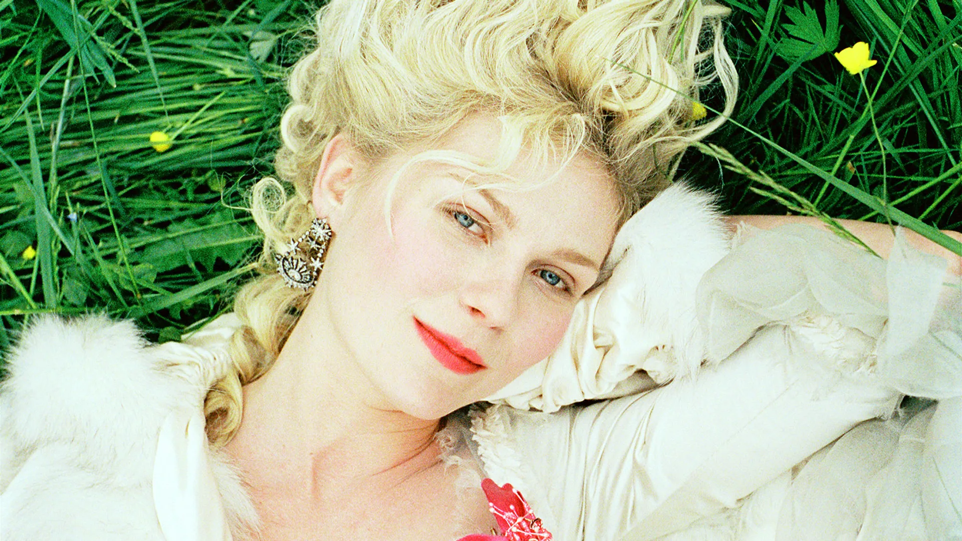 A still from the movie Marie Antoinette (2006) showing Kirsten Dunst lying on grass looking directly at the camera with her hand held up to her cheek, wearing white. She has red lips and some pink is showing on her dress.