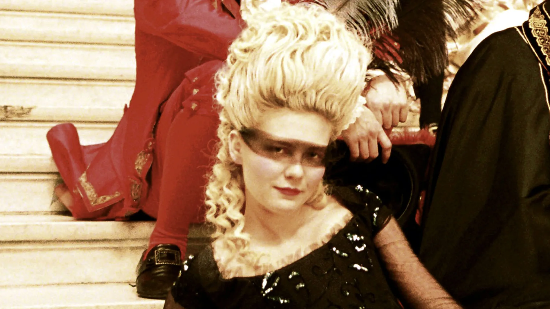 A still from the movie Marie Antoinette showing Kirsten Dunst dressed in a black dress with a see-through black band over her eyes. She is looking directly at the camera with a half smile as someone in red sits behind her and someone in black sits beside her on cream staircase.