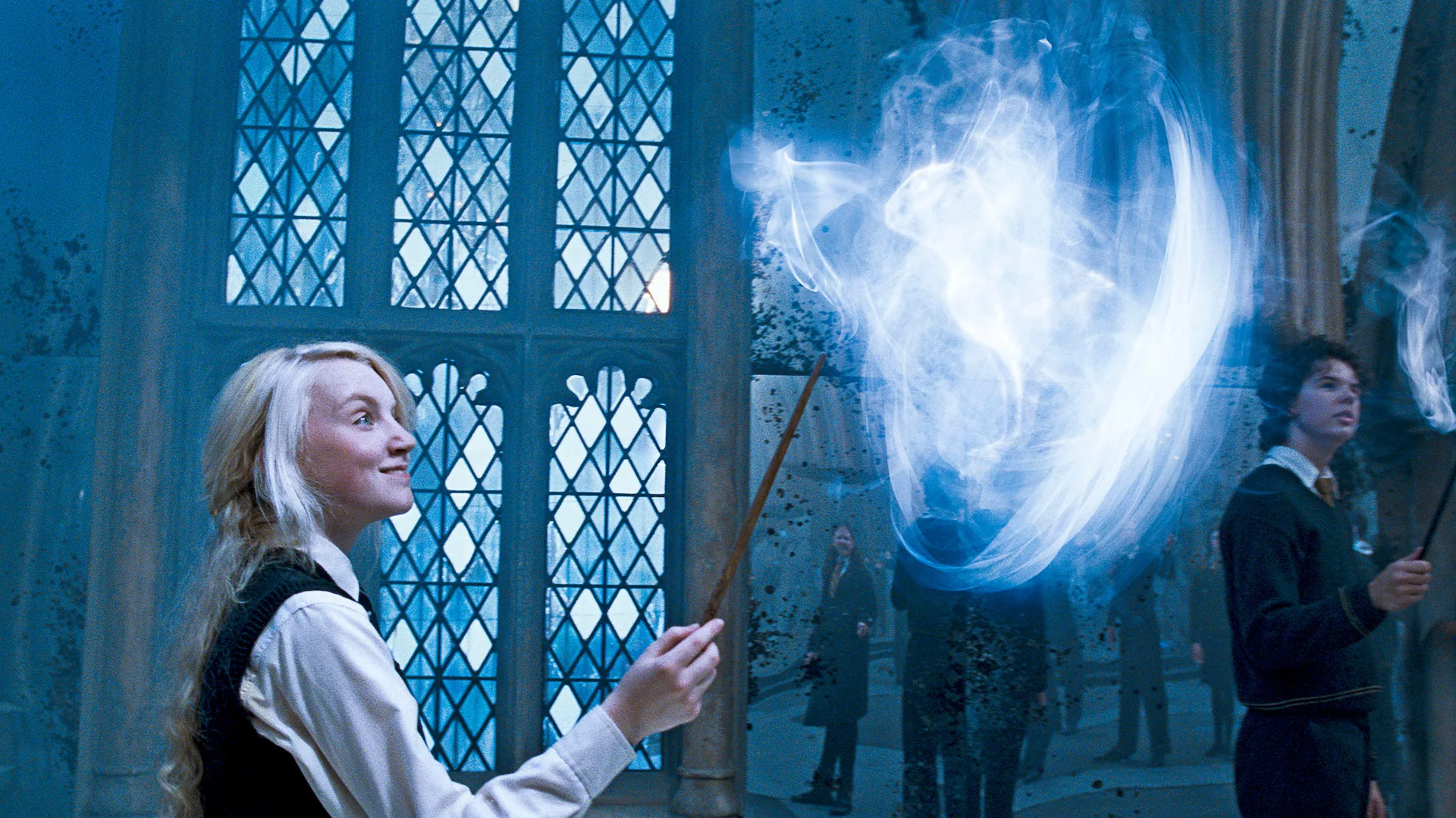 A still from Harry Potter movie showing Luna doing a spell with a bright white light coming from her wand. She is looking up at it smiling and behind her is a glass window.