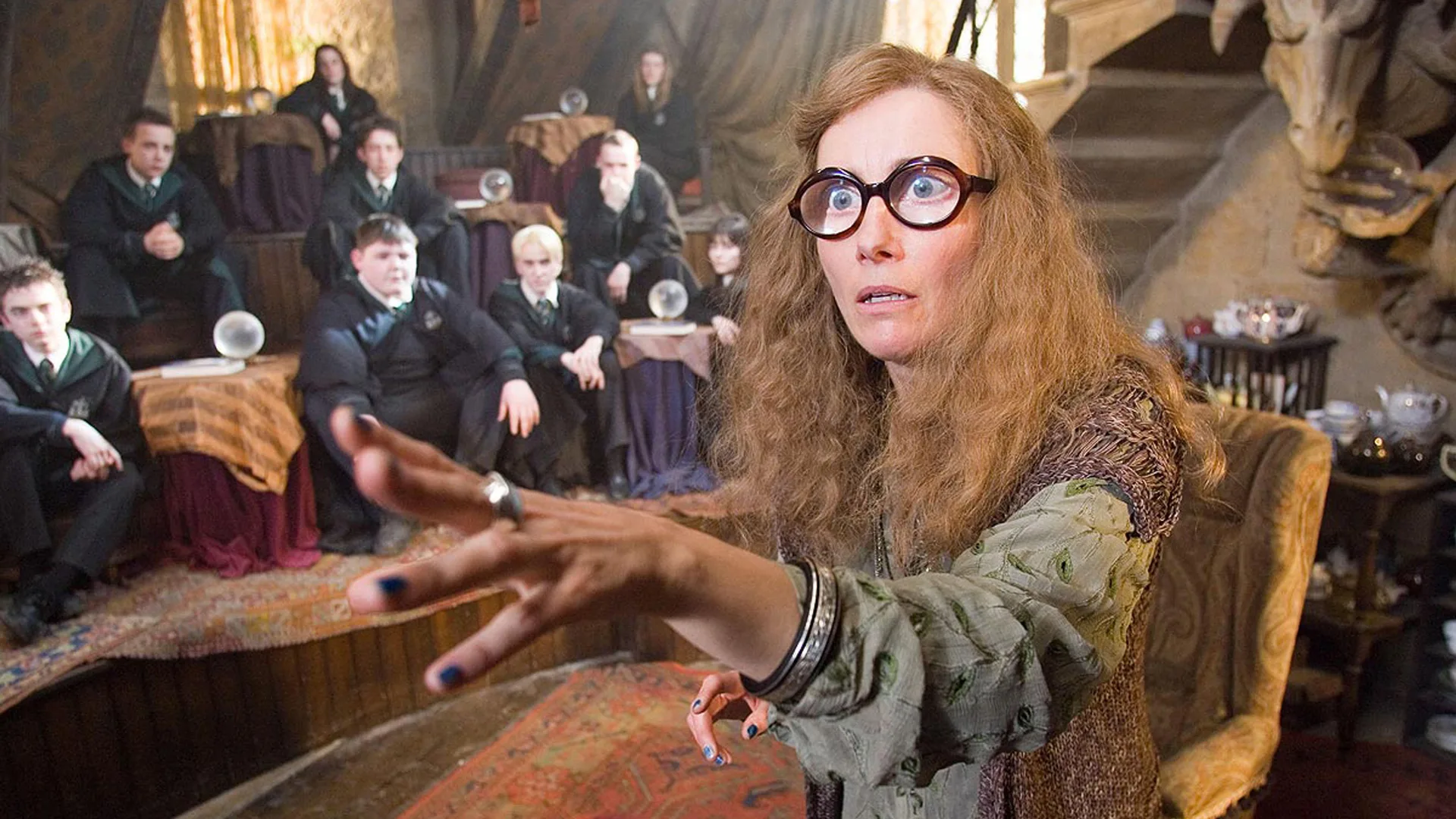 A still from Harry Potter showing Emma Thompson as Sybill Trelawney showing her in glasses with thick lenses holdiong her hand out to something in the distance as a class of students sit behind her watching.