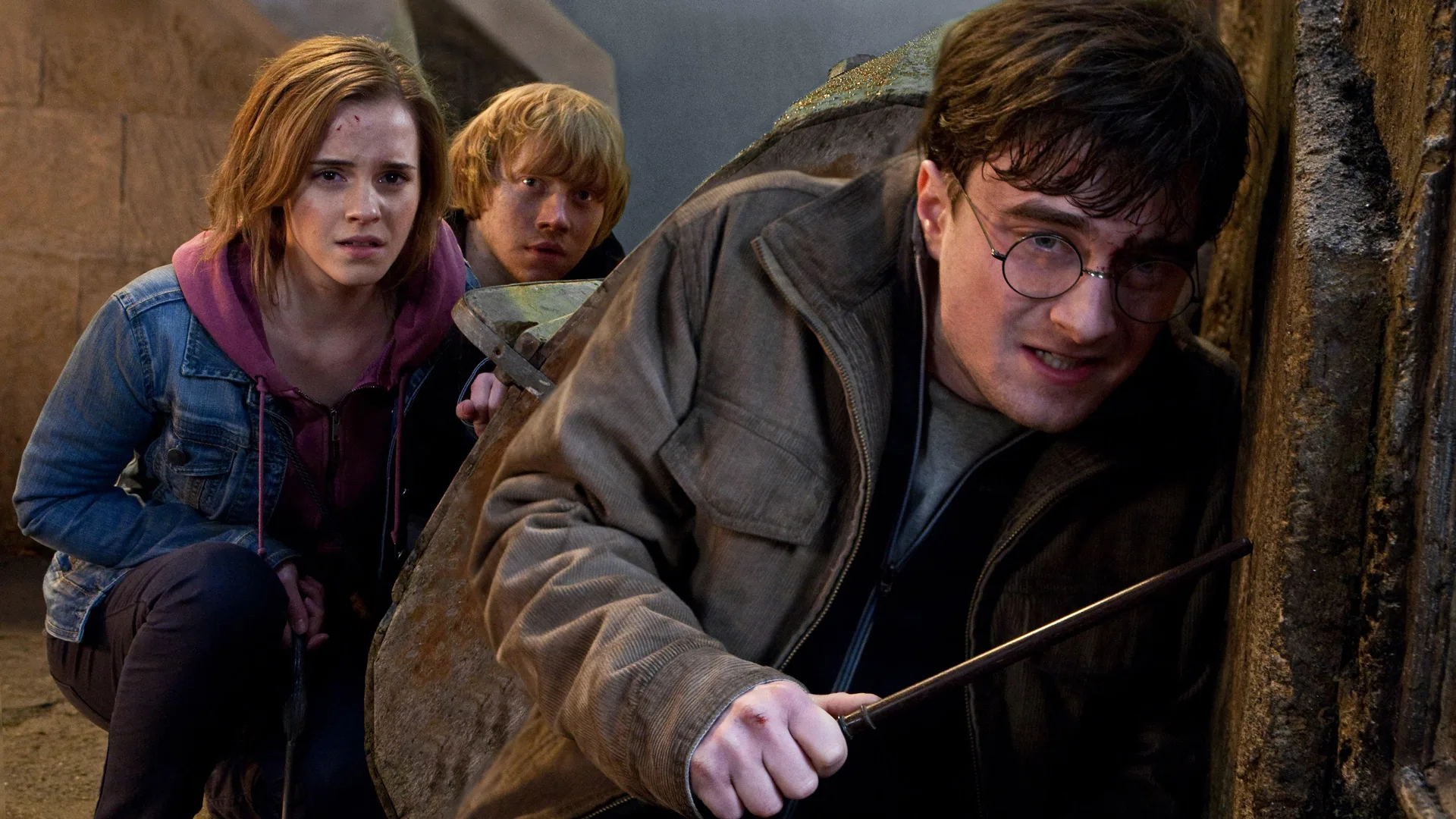 A still from Harry Potter and the Deathly Hallows Part 2 showing Harry, Ron and Hermoine hiding against a wall looking distressed. Harry is holding a wand.