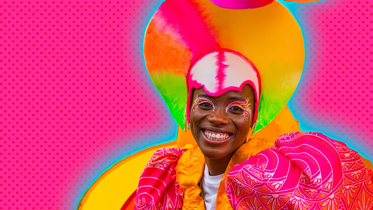 An image of a woman wearing a bright pink, orange and yellow outfit for carnival with a circular headdress and giant shoulders with decorative silver and orange floral motifs. She is wearing pink, orange and white eye makeup and smiling directly at the camera. She is set against a pink dotted background with a blue halo.