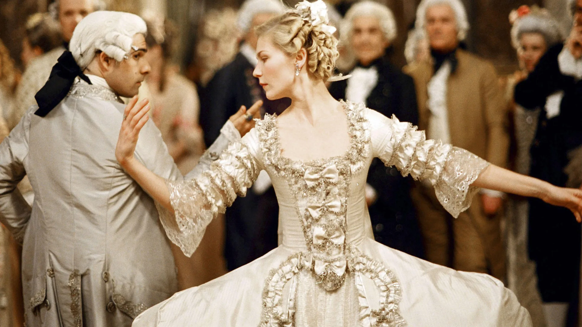 A still from the Sofia Coppola move Marie Antoinette (2006) showing Kirsten Dunst as Marie Antoinette dancing with her husband King Louis - she is wearing a grey beige dress with frills and turning her head to the side holding her hand out to her husband. There are people behind her in the background.