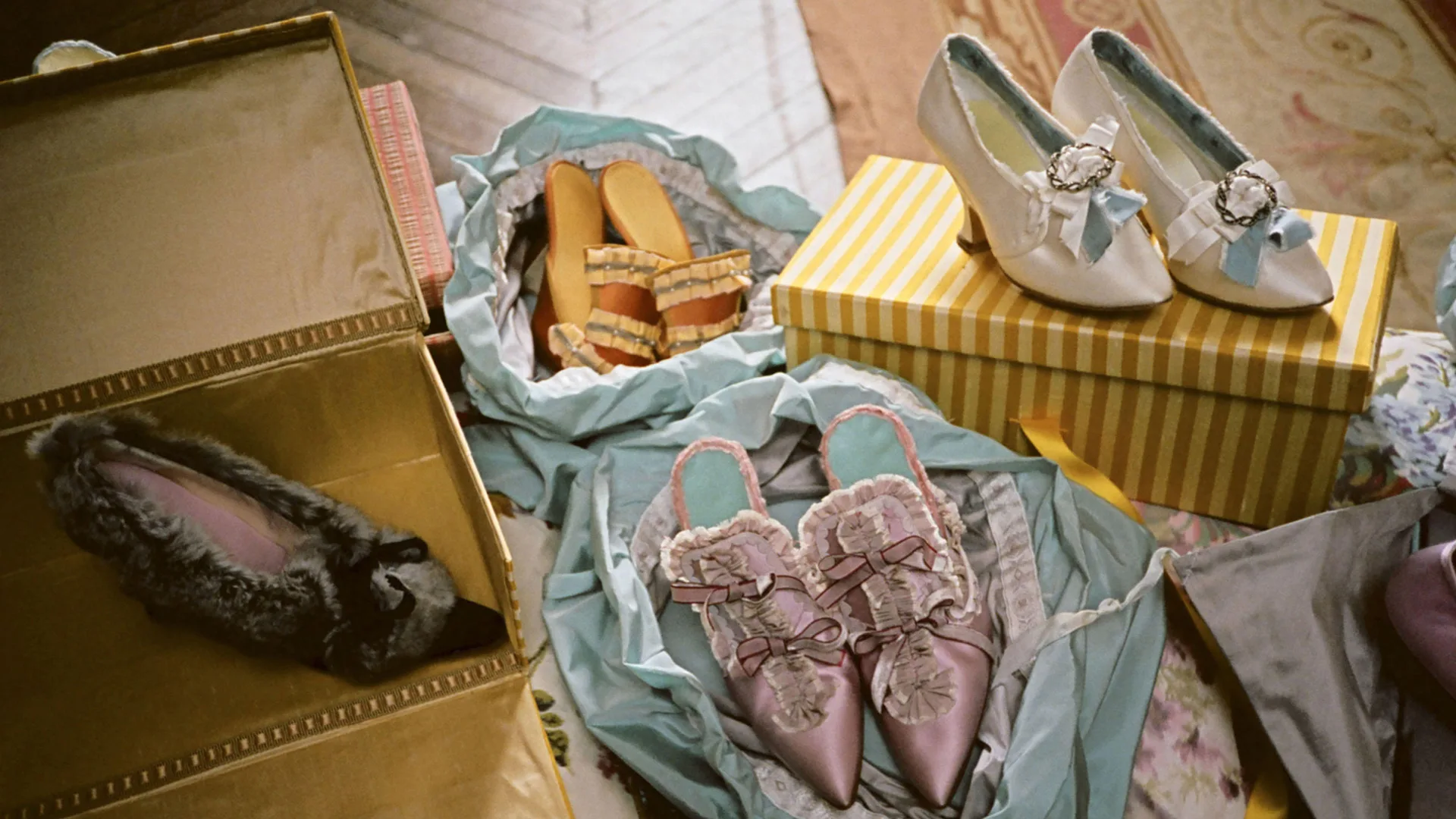 A photo of some Rococo shoes in pairs placed on blue satin surrounded by yellow striped boxes.