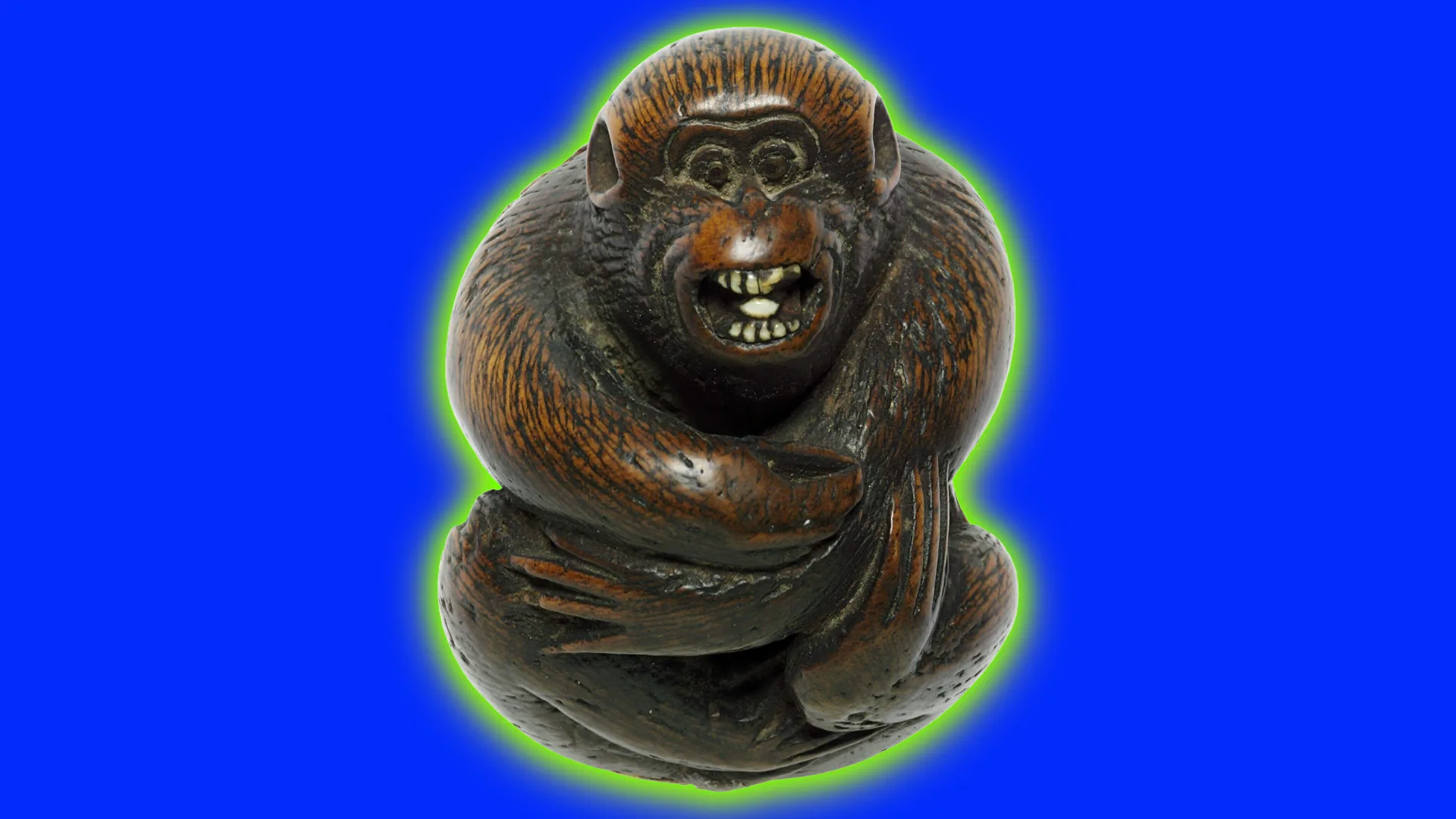 An image of a Netuske monkey sculpture made out of stag antler showing a monkey in a sitting pose with its arms wrapped around it and mouth open. It is set against a blue cobalt background with a green halo.