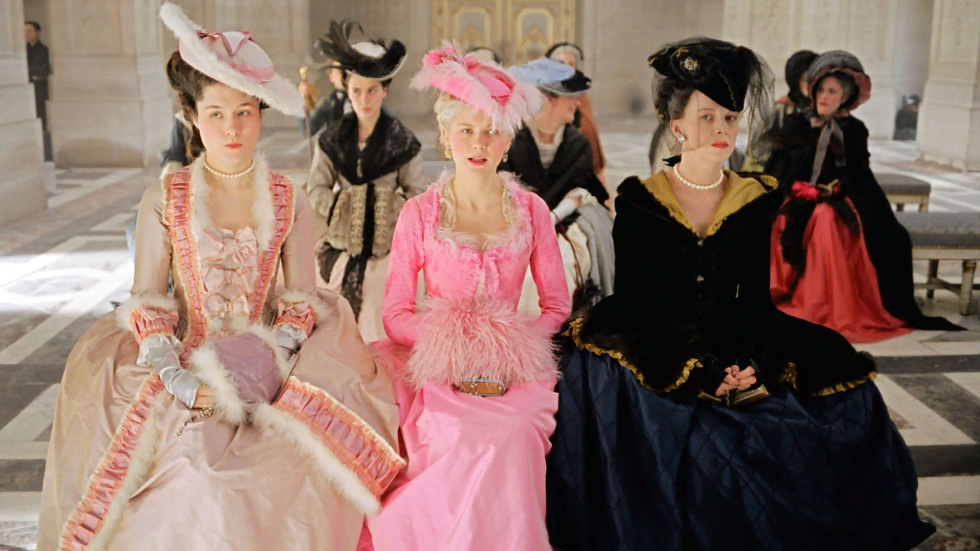 A still from the Sofia Coppola move Marie Antoinette (2006) showing Kirsten Dunst as Marie Antoinette sat with two women either side of her. She is wearing a bright pink dress and hat with her hands on her lap. The woman to her left is wearing all black and the woman to her right is wearing a cream and pink dress. They're in a room with other people with a black and white floor.