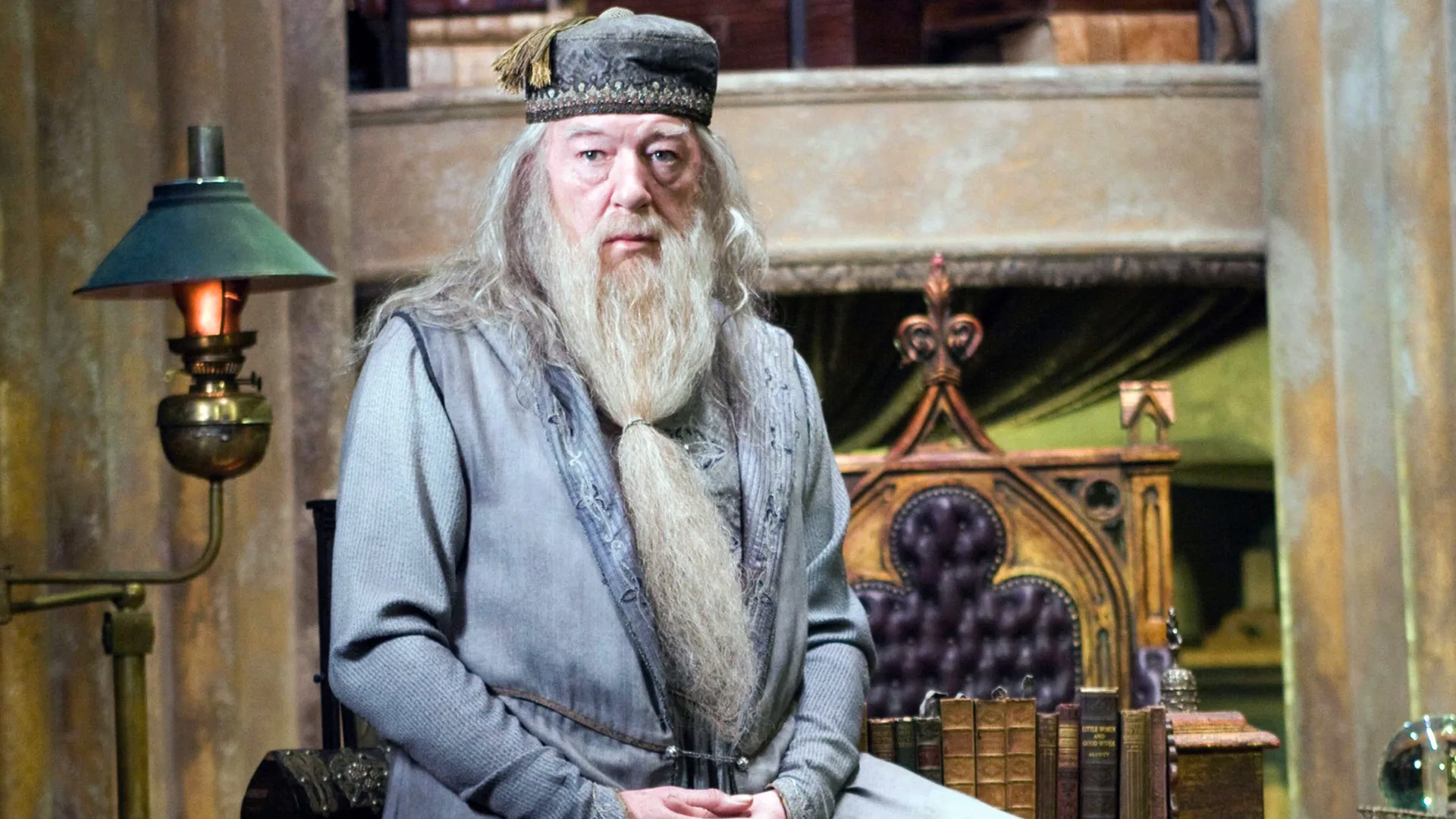 A still from Harry Potter showing Michael Gambon as Dumbedore sat on a share staring off to the distance wearing a grey cloak and hat.