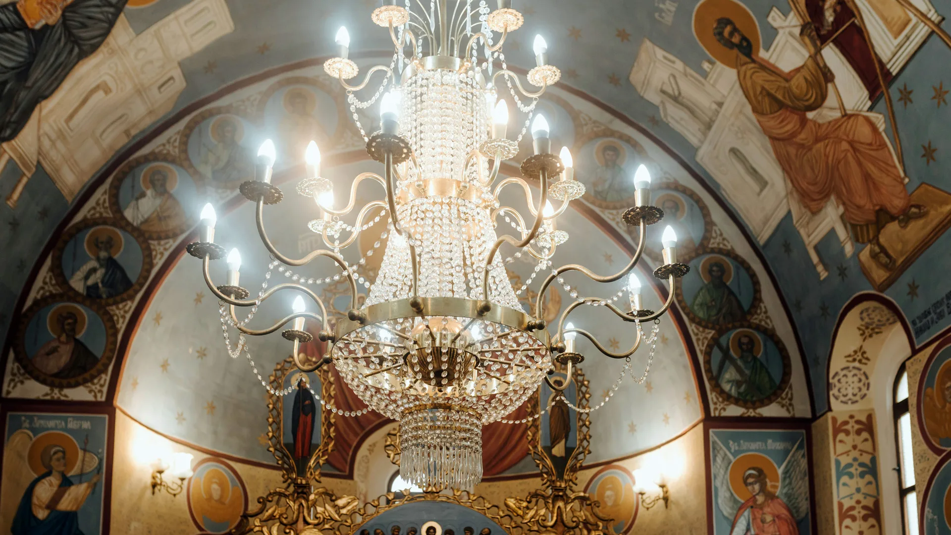 A photo of a crystal chandelier hanging from a painted ceiling with bright shining bulbs.