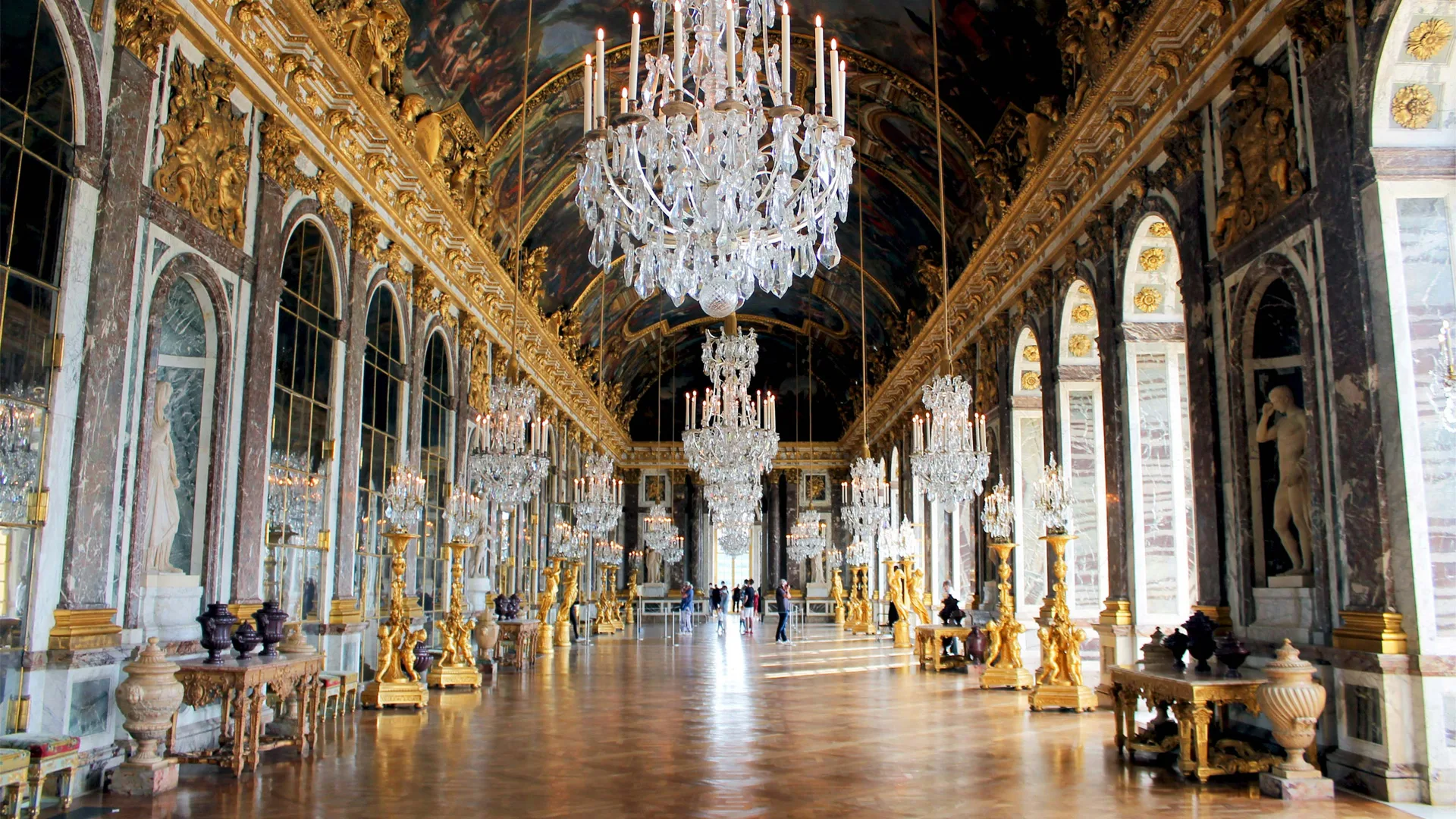 A photo of the hall of mirrors inside Versailles showing a grand room with mirrors either side of the parkor flooring and a giant crystal chandelier hanging from the painted ceiling.