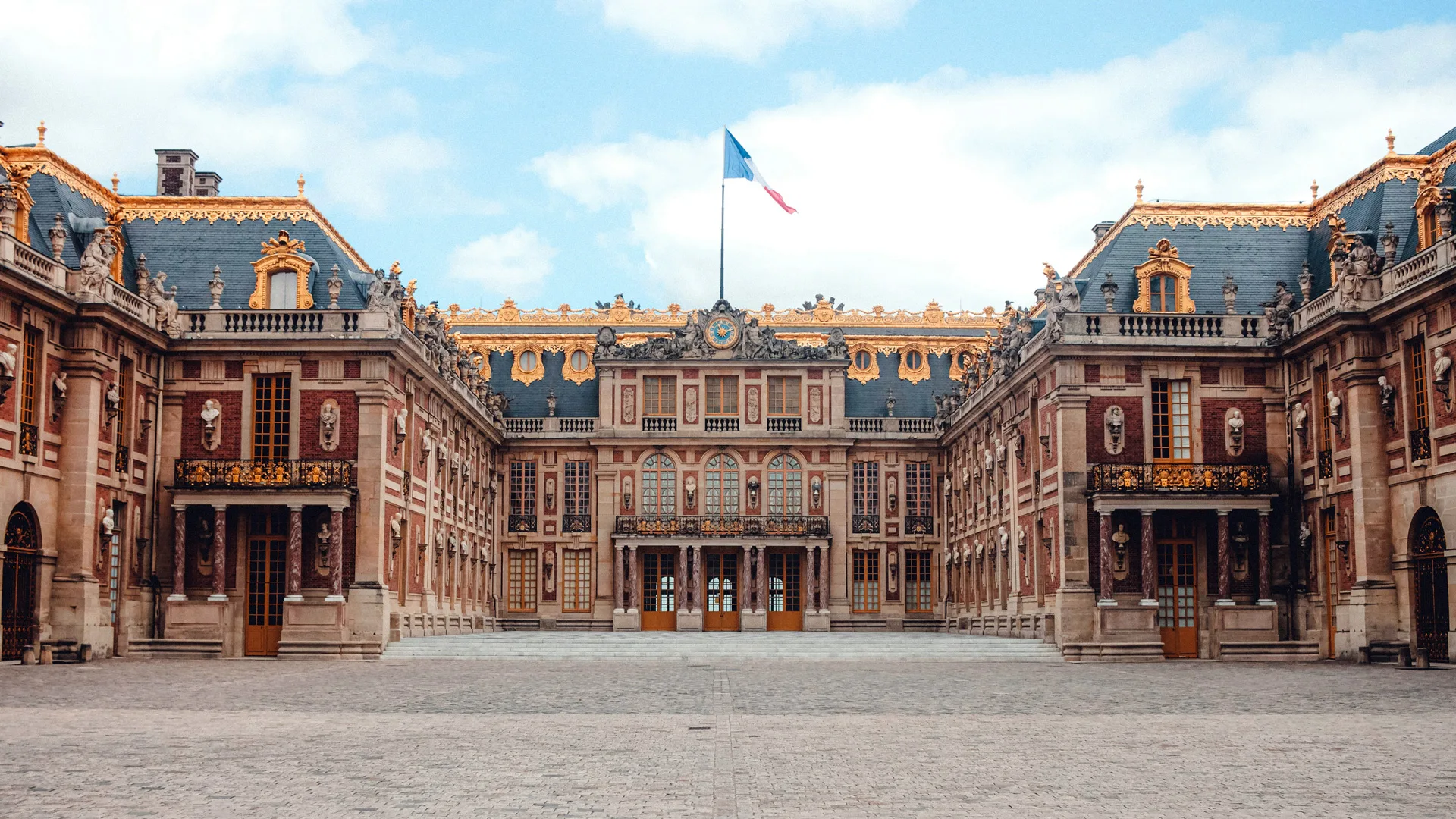 A photo of the facade Versailles showing the main courtyard and pink and blue gilded building with a flag flying in the central roof and blue cloudy sky above.