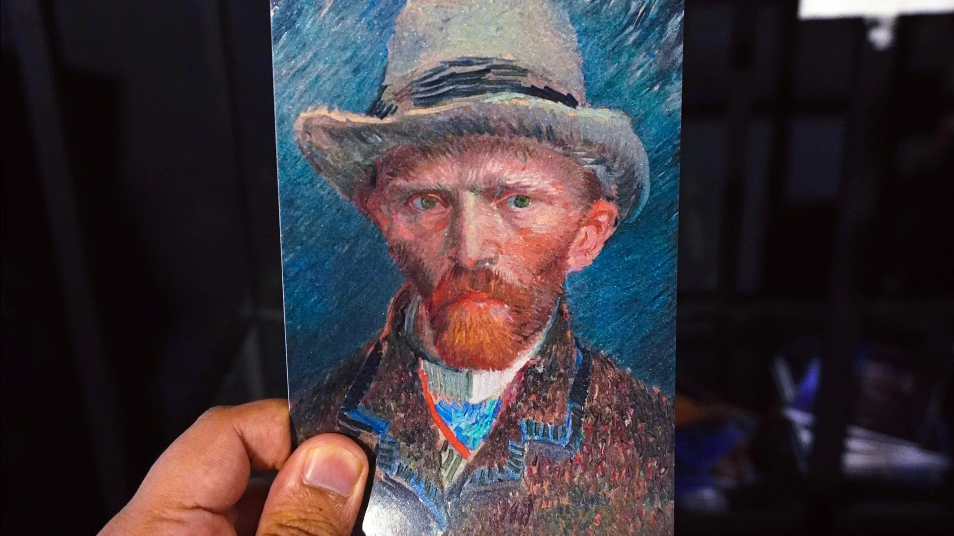 A painting postcard of artist Van Gogh showing him wearing a hat, shirt and tie and brown jacket against a blue background. The postcard is being held by a hand against a black background.
