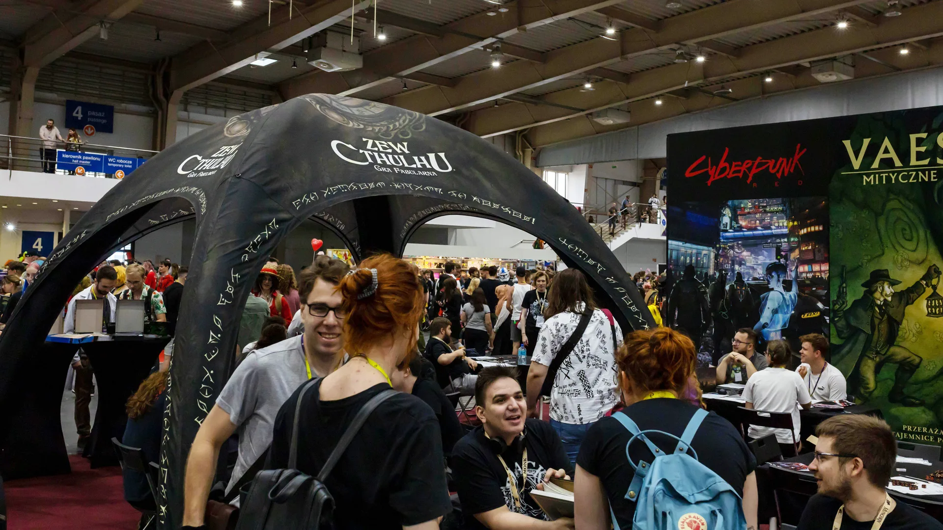 A photo of a games convention with a gazebo showing "Call of Cthulhu" in Polish and people underneath all talking and engaging over the game.