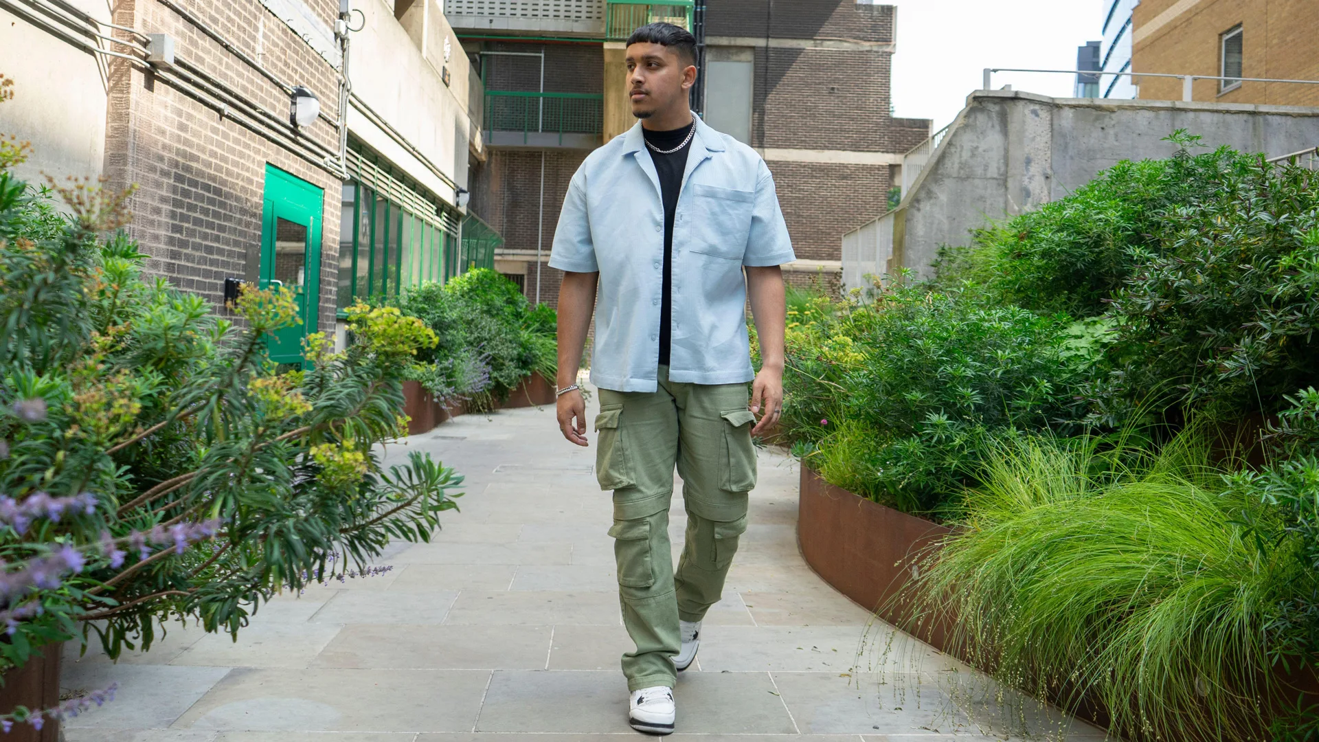 A photo of a man wearing a blue shirt and green cargo pants and white trainers walking through a block of flats with green foliage either side.