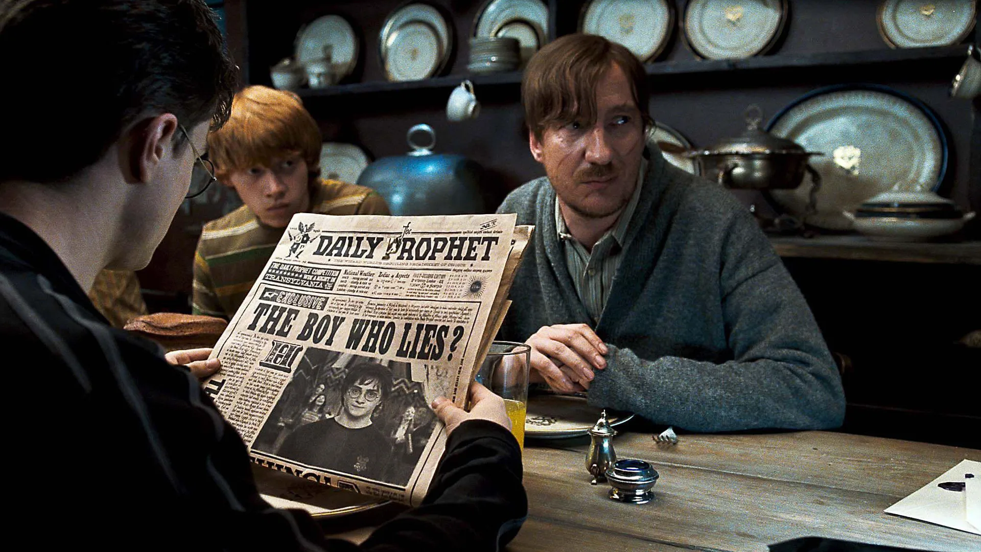 A still from Harry Potter and the Order of the Phoenix showing David Thewlis as Remus Lupin sat at a table looking concerned in a blue grey jumper whilst Harry sits opposite him reading a newspaper with the headline "THE BOY WHO LIES" and Ron sat next to Remus with a sombre expression. There are plates on a cabinet behind them.