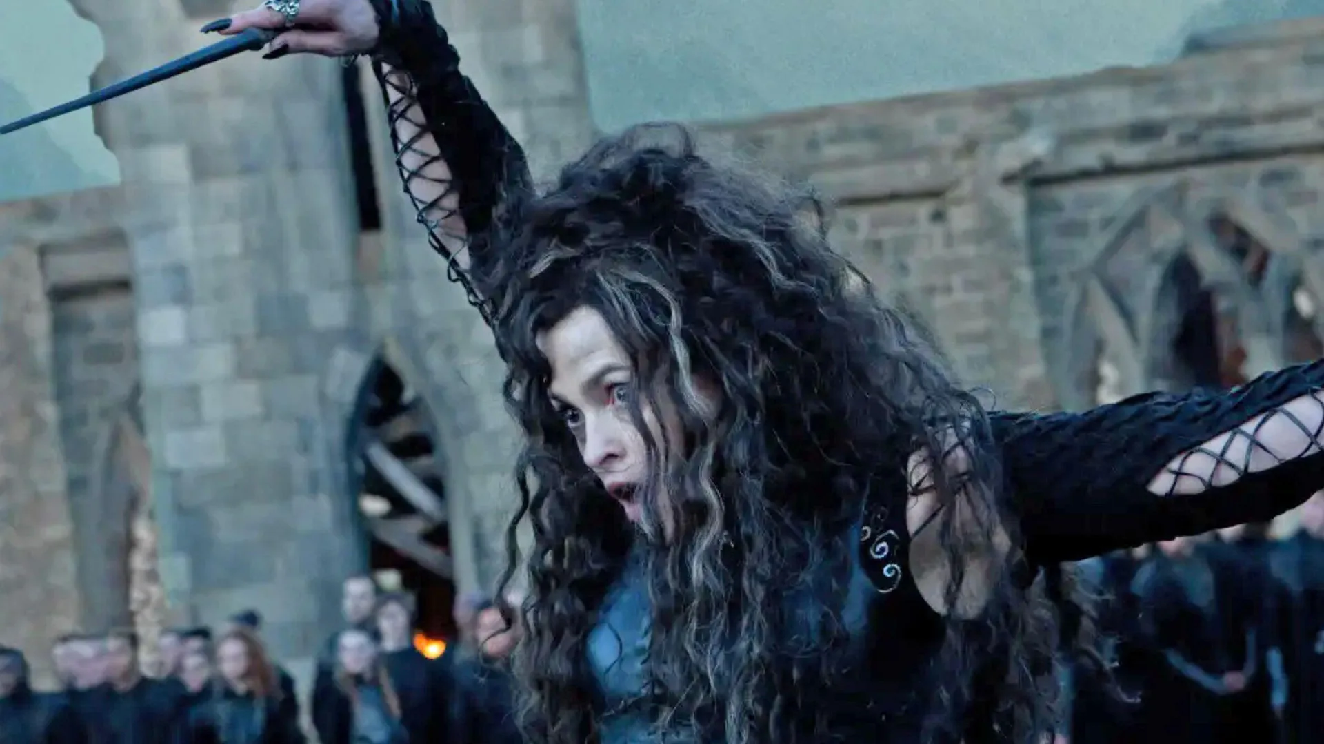 A still from Harry Potter and the Deathly Hallows Part 2 showing Bellatrix LeStrange holding up her wand with an open mouth as her hair covers her face against a bricked building and crowd watching.