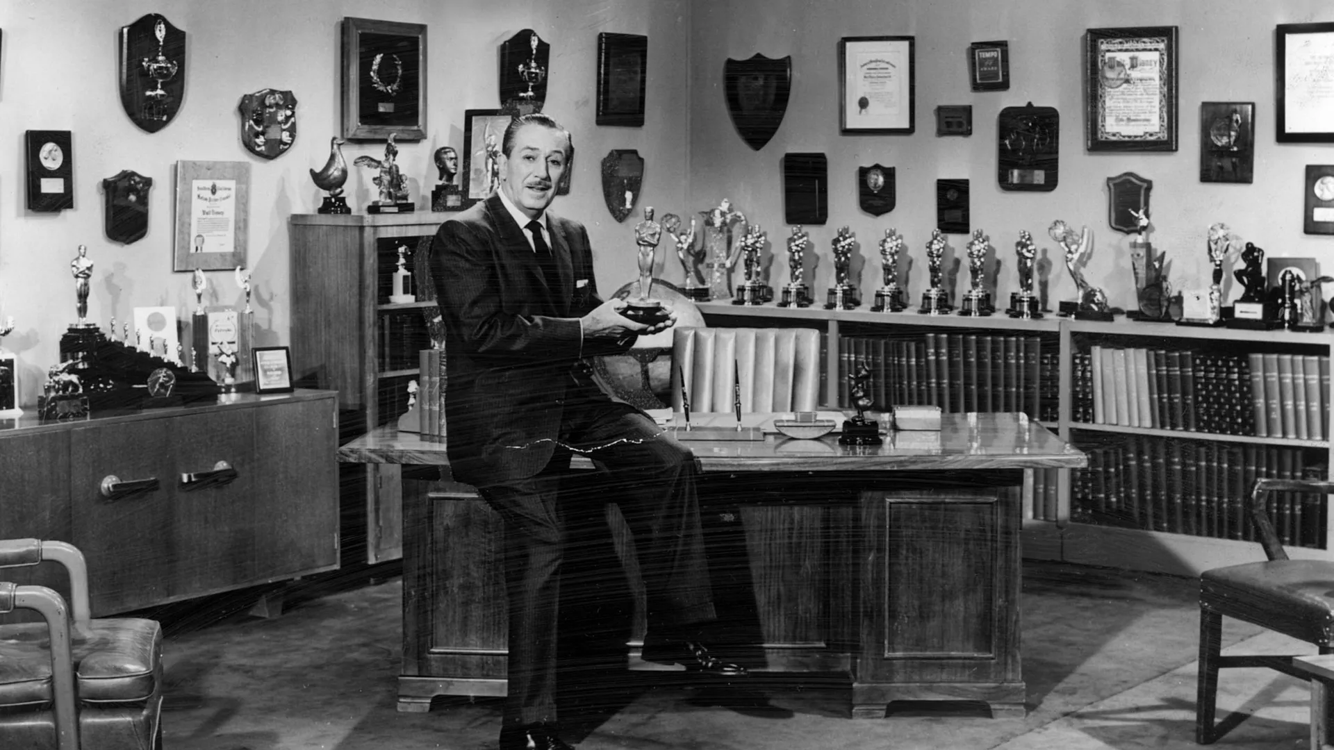 A black and white photo of Walt Disney sat in his office holding his Oscars whilst sat on a chair beside a desk with more trophies and awards on the wall and shelf behind him.