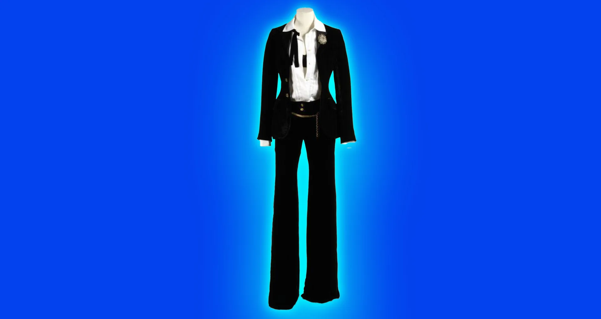A photo of a V&A collection of a Dolce and Gabanna women's suit with black trousers, white shirt and a bow tie undone hanging from the mannequin's neck. It is set against a blue background with light blue halo.