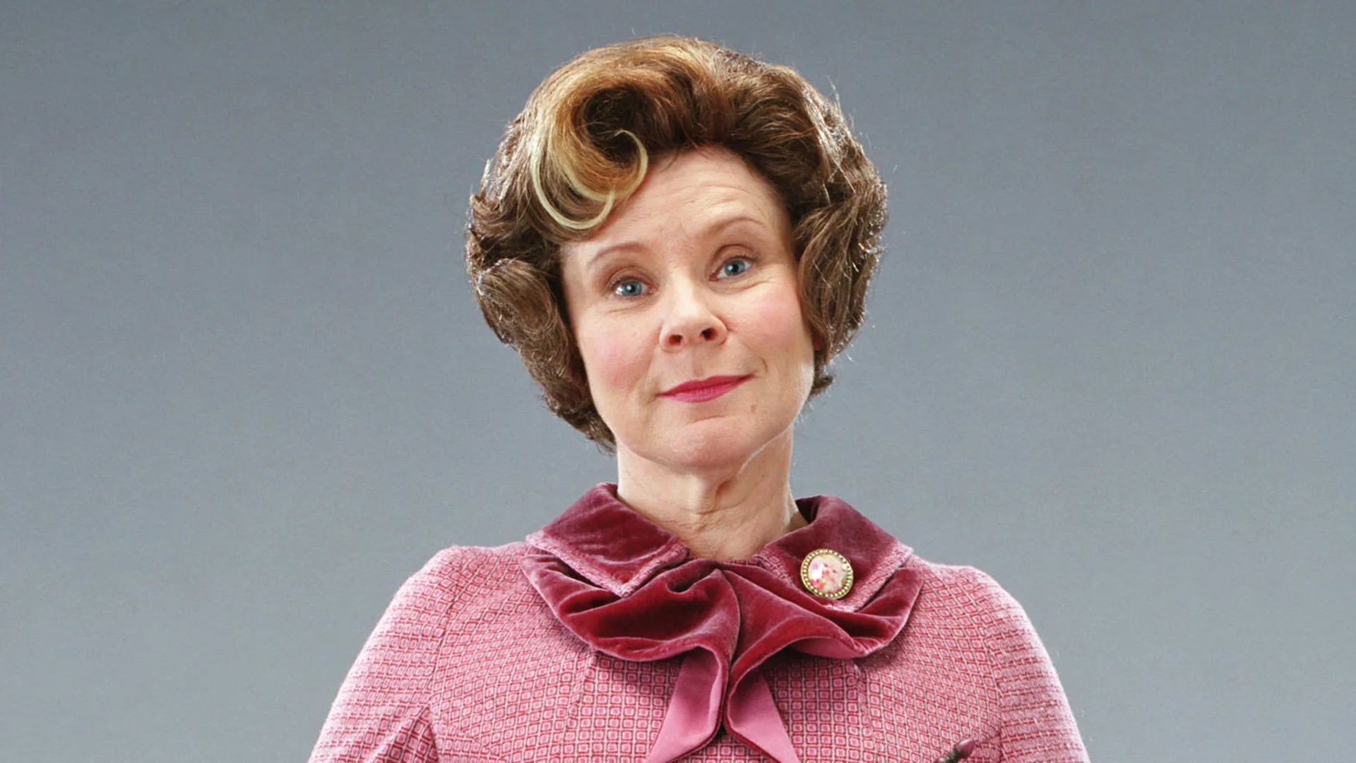 An image of Dolores Umbridge from Harry Potter stood against a grey background in her pink outfit half-smiling directly at the viewer.