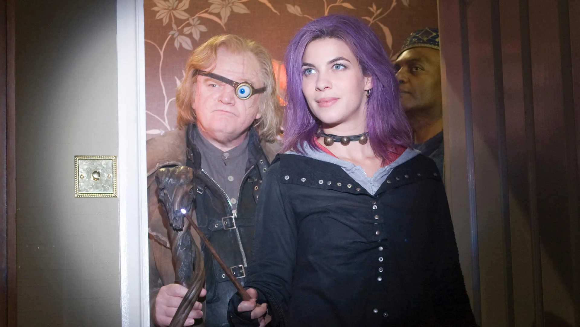 An image of Professor Moody and Tonks stood in a door way of a house with a spotlight shone on them looking off to something in the room. Moody has a fake eyeball in a patch on his face and Tonks has purple hair and a black jacket.