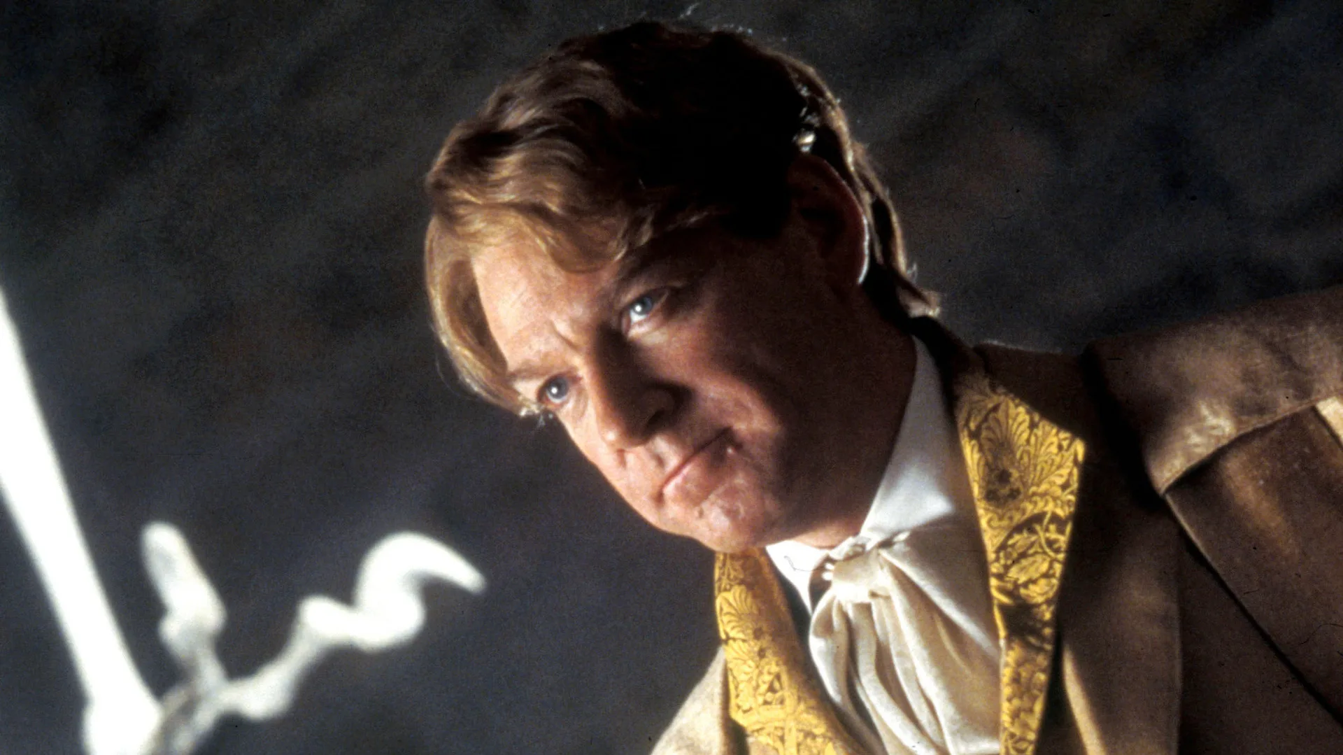 A still from Harry Potter and the Chamber of Secrets showing Kenneth Brannagh as Professor Lockhart wearing a gold jacket and white shirt half smiling to the side with a black background and white light to his left.