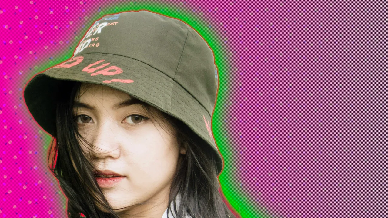 A photo of a girl wearing a green bucket hat with dark hair slightly over her face against a pink dotted background with a green halo.