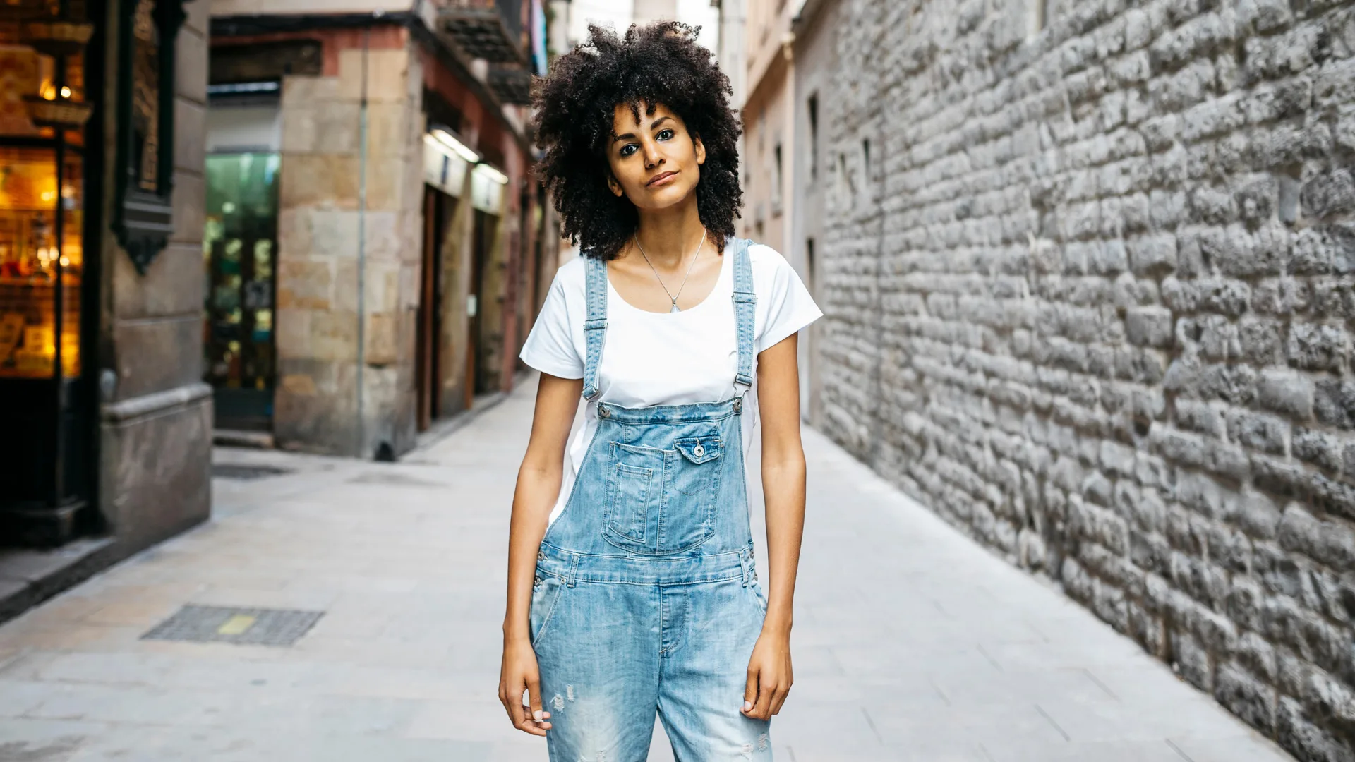 A photo of a woman wearing blue dungarees and a white tshirt stood in a street with a brick wall one side and a restaurant window with warm lights the other side.