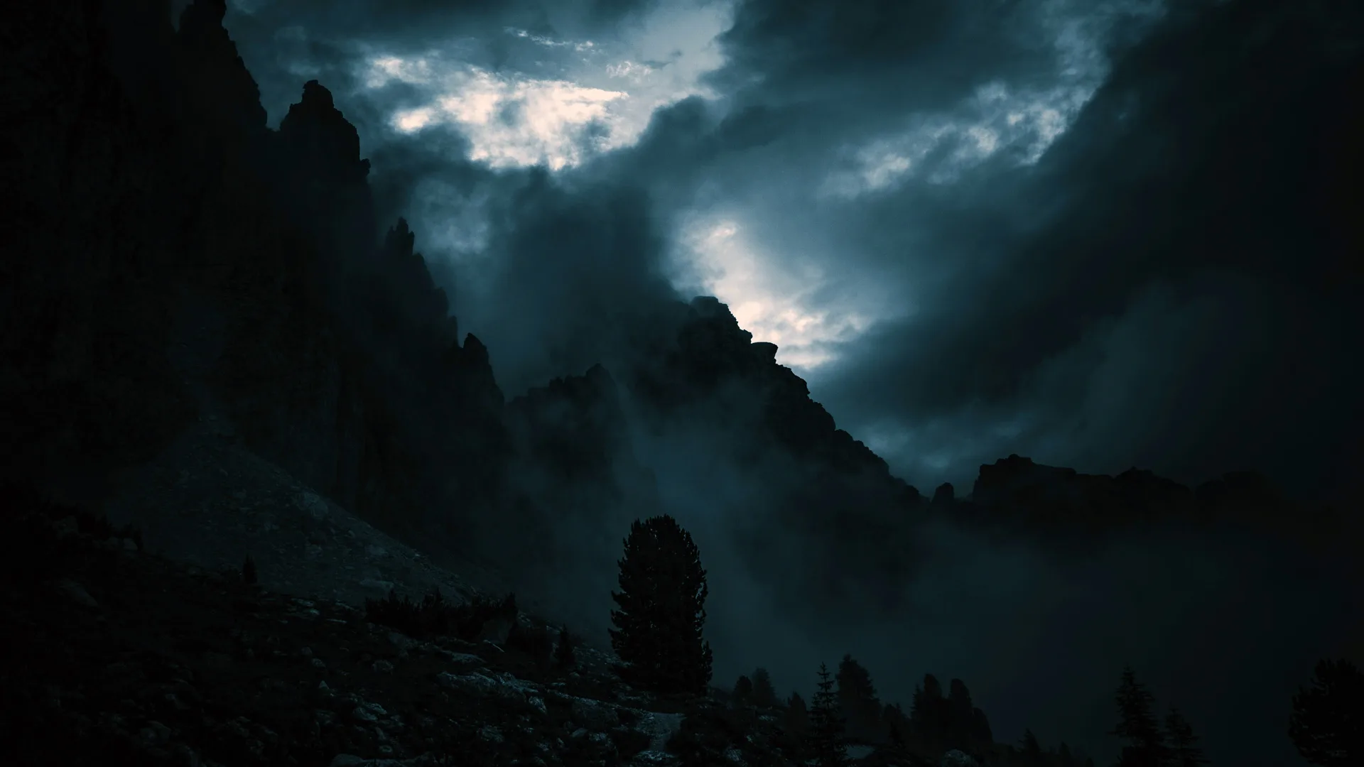 An image of a mountain against a stormy moody sky. The picture is almost black except for some of the light coming behind the clouds.