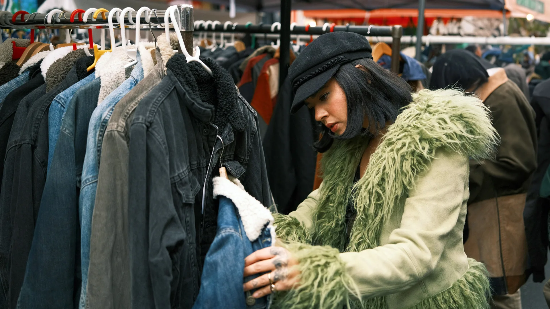 A photo of a woman wearing a green afghan style coat with black hat holding a denim jacket in her hands with a rail of coats hanging beside her.