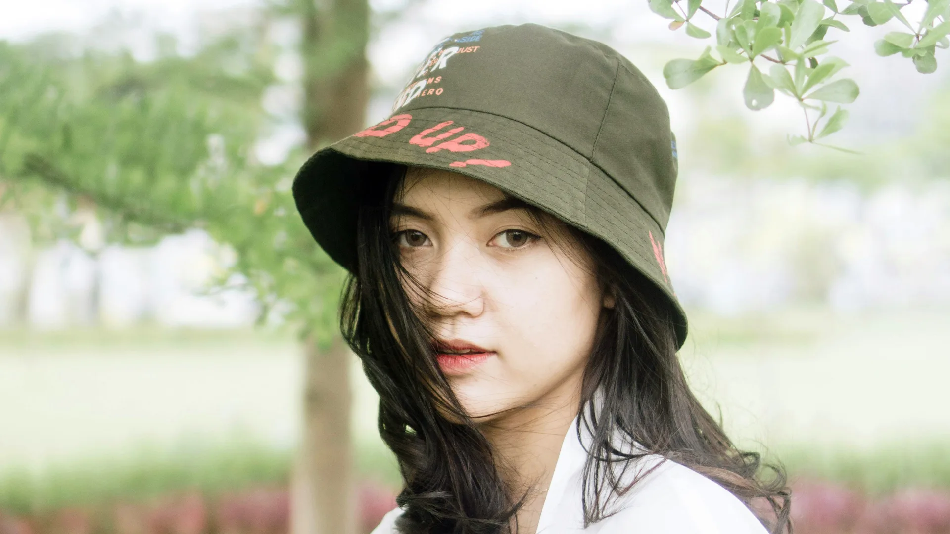 A photo of a young woman wearing a green bucket hat. She has dark hair that is slightly over her face. She is stood against a tree and park background.