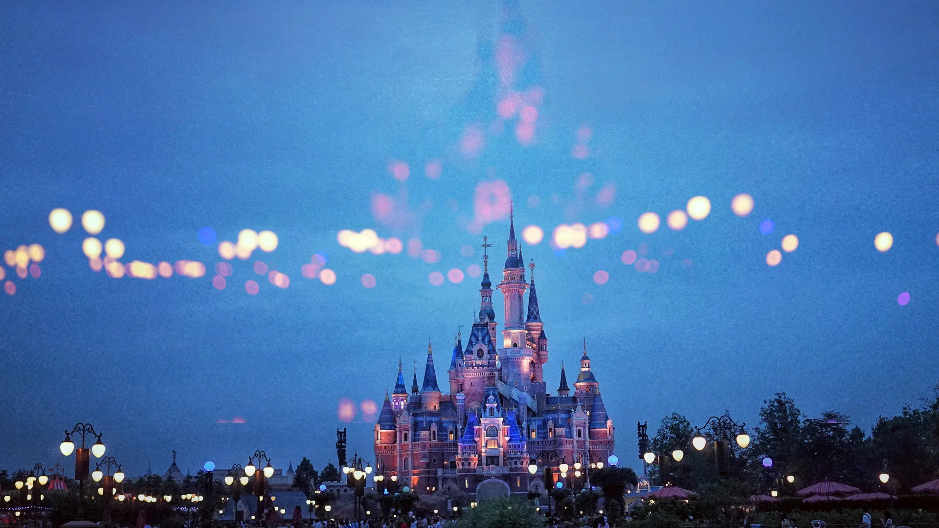 A photo of Cinderella's castle from a distance at dusk showing the castle glowing pink and purple with bokha lights across the castle and below.