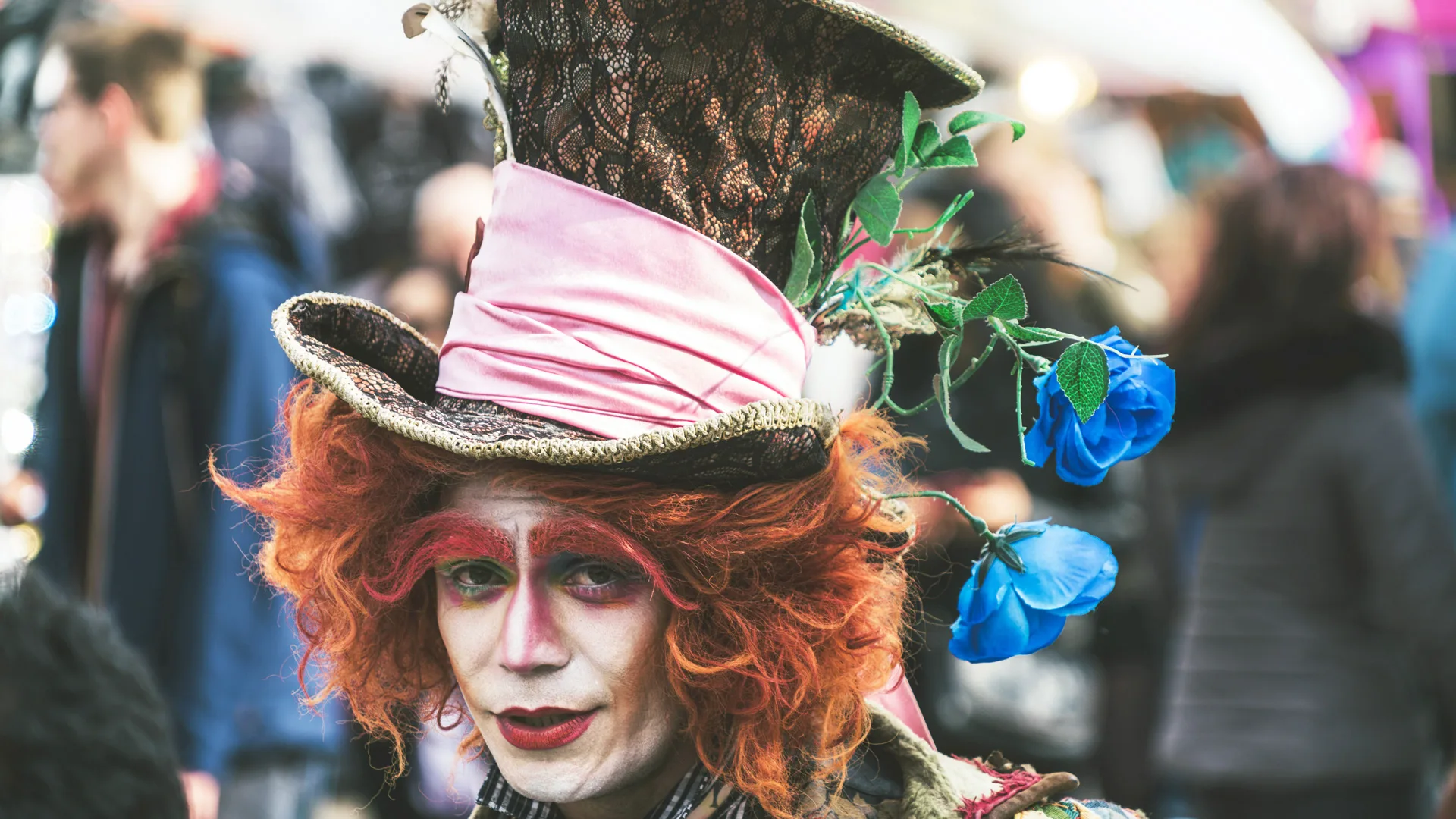 A photo of a person dressed as the Mad Hatter in an orange wig, eyebrows and a big green and pink top hat with white face makeup and red eyeshadow. There are also blue flowers hanging from the hat.