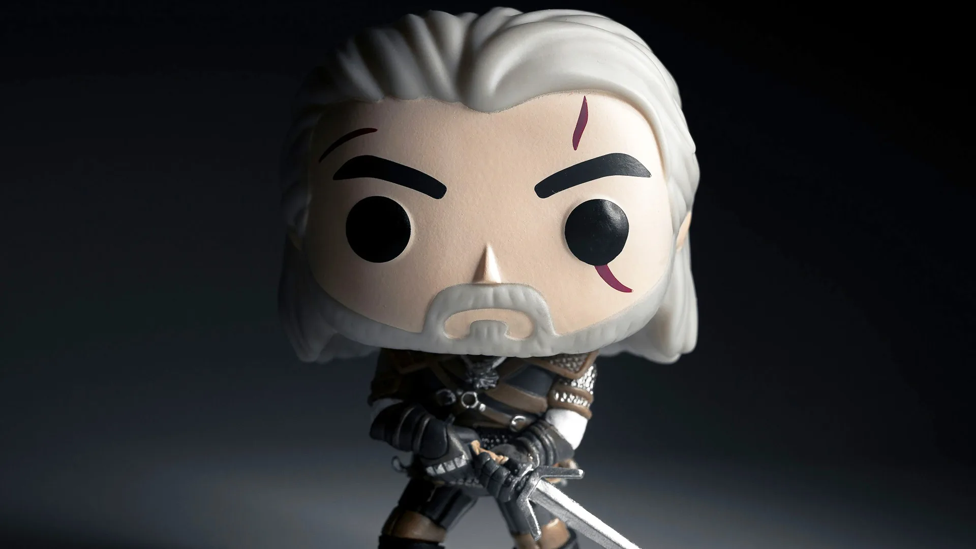 An image of The Witcher as a Funkopop toy with white hair, a scar over his left eye, a black outfit and he's holding a sword - stood against a grey and black background.