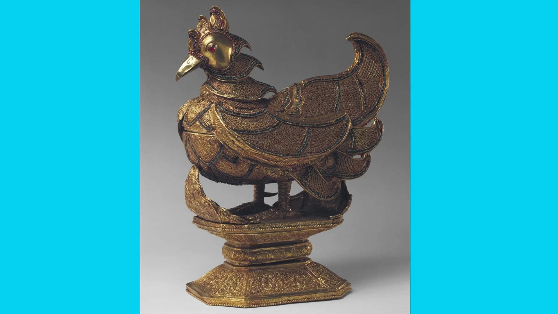 A photo of a gold and jewelled box in the shape of a bird stood on a plinth with blue borders either aside.