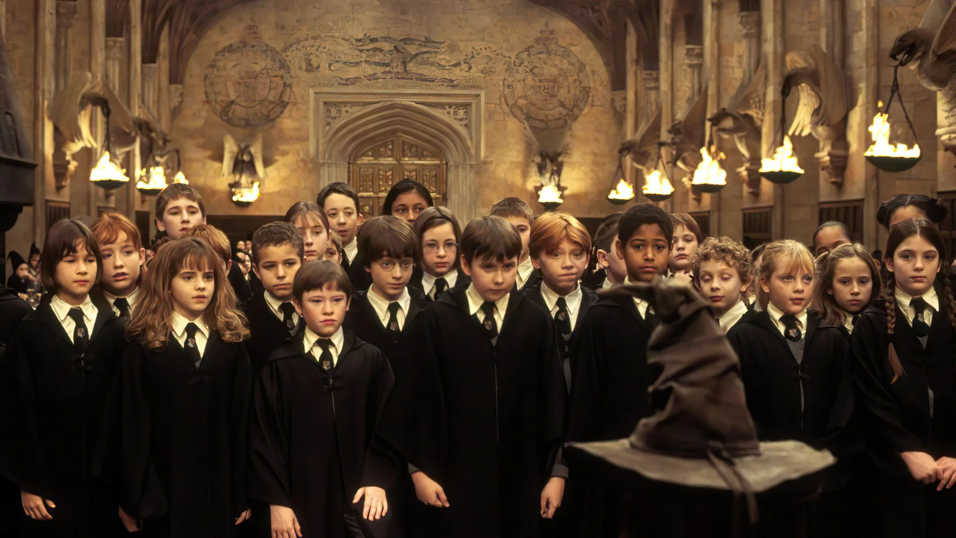 A still from Harry Potter and the Philopsher's Stone showing the children stood together in front of the sorting hat in the great hall with lights behind them.