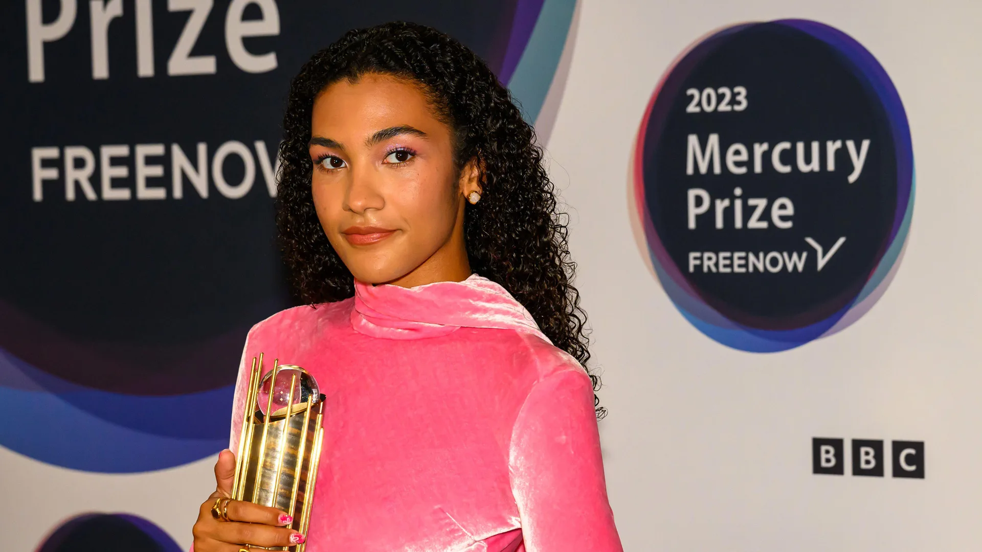 A photo of singer Olivia Dean wearing a pink velvet top holding a gold Mercury Prize looking off to the side with the Mercury Prizes logos on a wall behind her.
